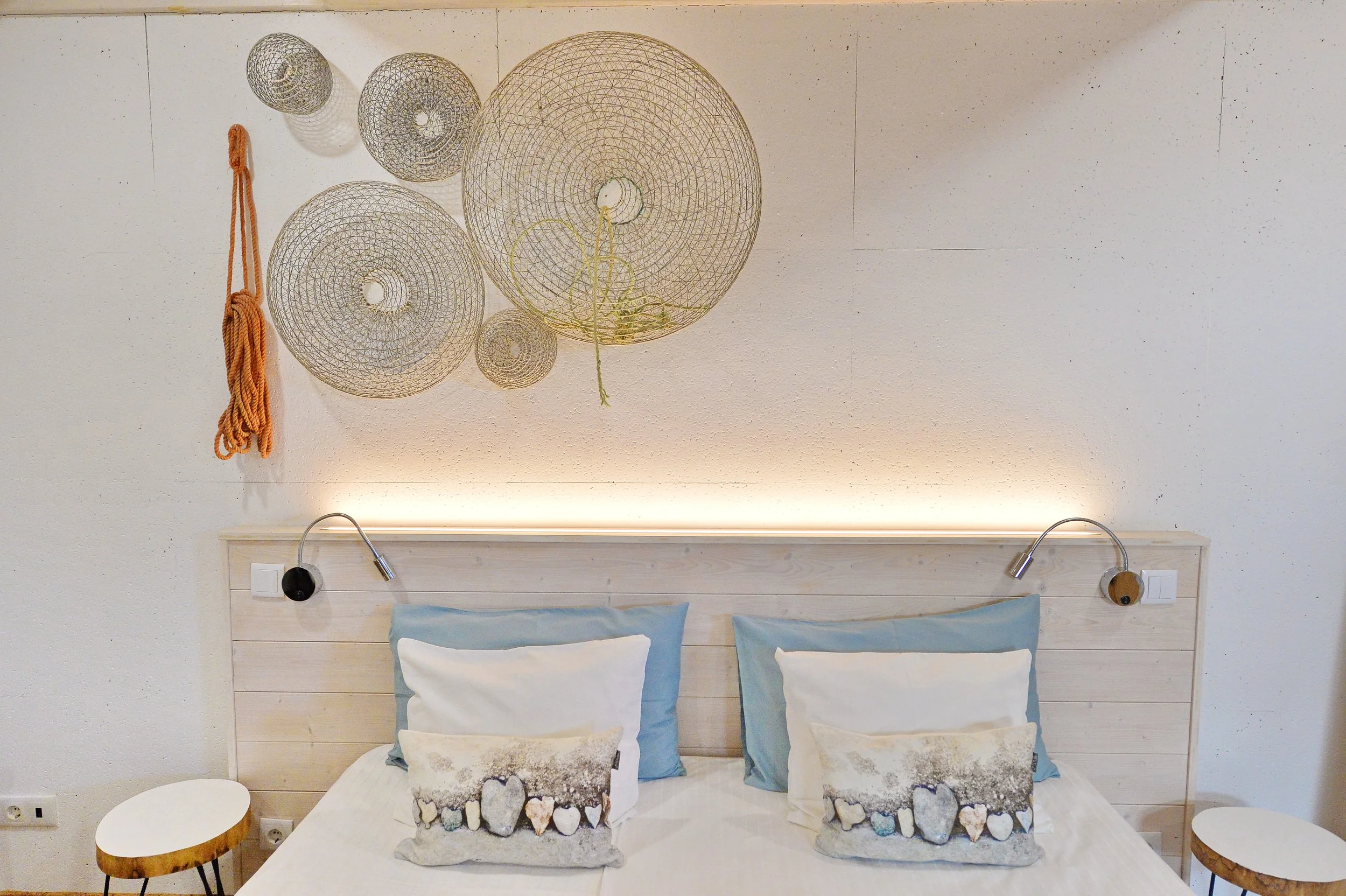 A bed with a light-colored wooden headboard, two pillows with light blue pillowcases, and two decorative pillows with stone patterns. Above the bed, there are decorative wire spheres and a bundle of orange rope mounted on a white wall. Two adjustable reading lights are attached to the headboard on either side of the bed. There are two small, round wooden side tables, one on each side of the bed.