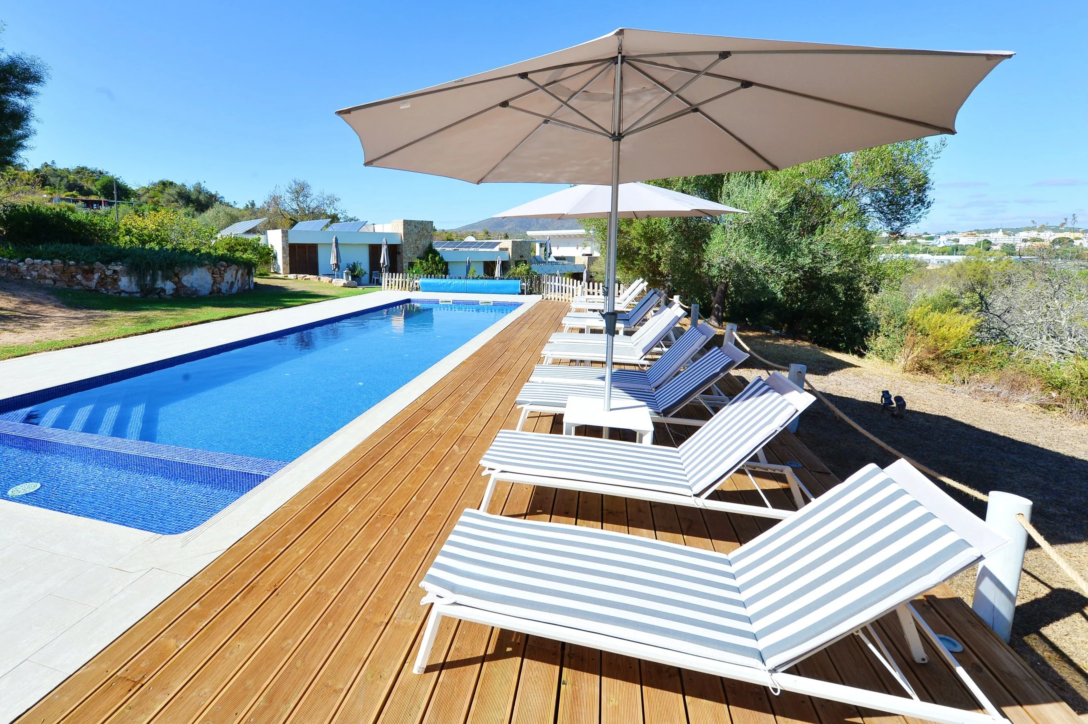 Empty poolside area with striped lounge chairs, large umbrellas, and a wooden deck, overlooking greenery and clear blue sky.