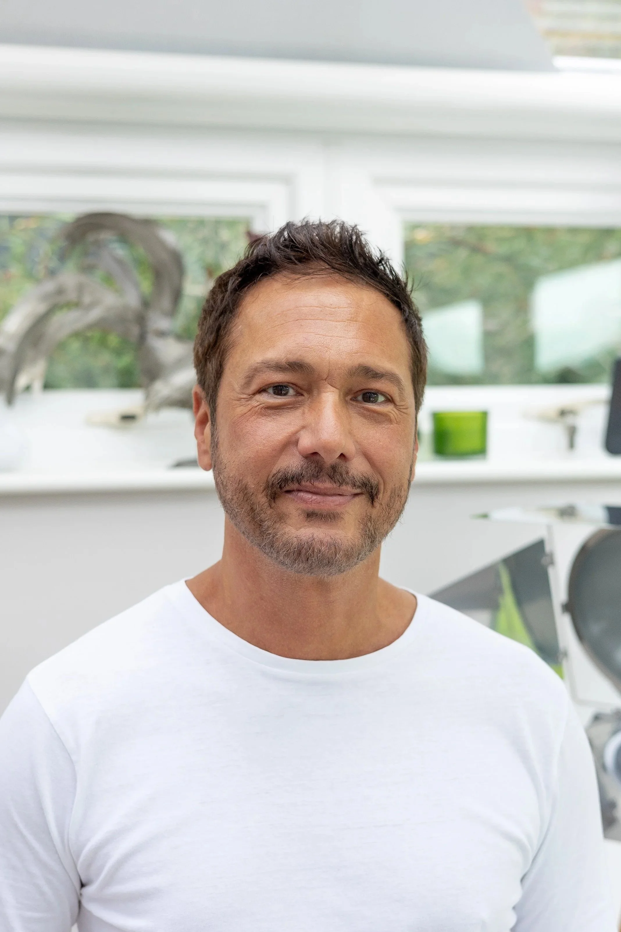 A man with dark hair, a beard, and a mustache, wearing a white shirt, smiling in a bright room with large windows and art sculptures in the background.