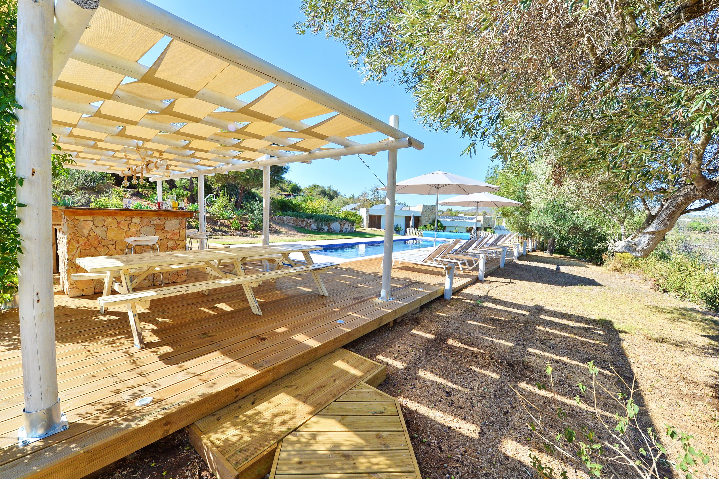 Outdoor swimming pool area with lounge chairs, umbrellas, and a shaded wooden deck surrounded by trees and greenery
