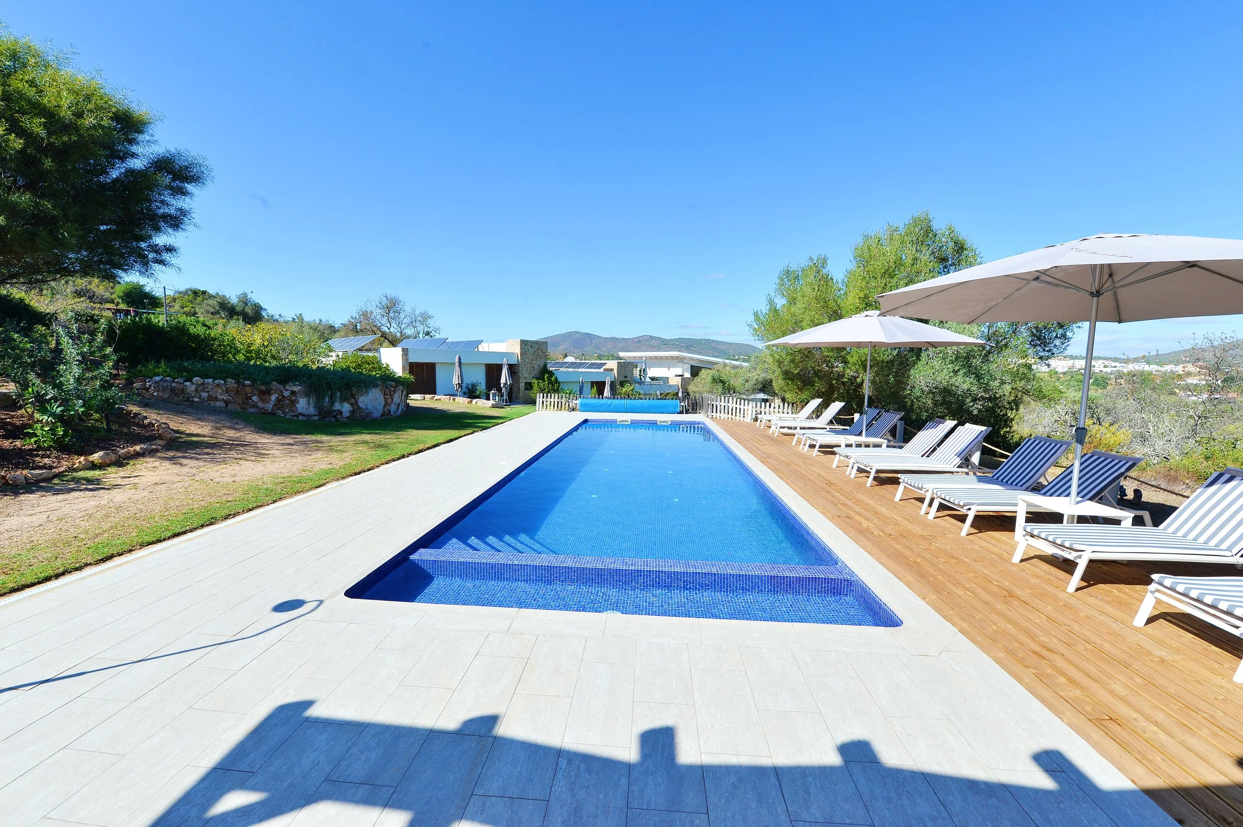 A modern swimming pool with blue tiles is surrounded by a white tiled deck and a wooden lounging area with white sunbeds and umbrellas. Green trees and hills are in the background under a clear blue sky.