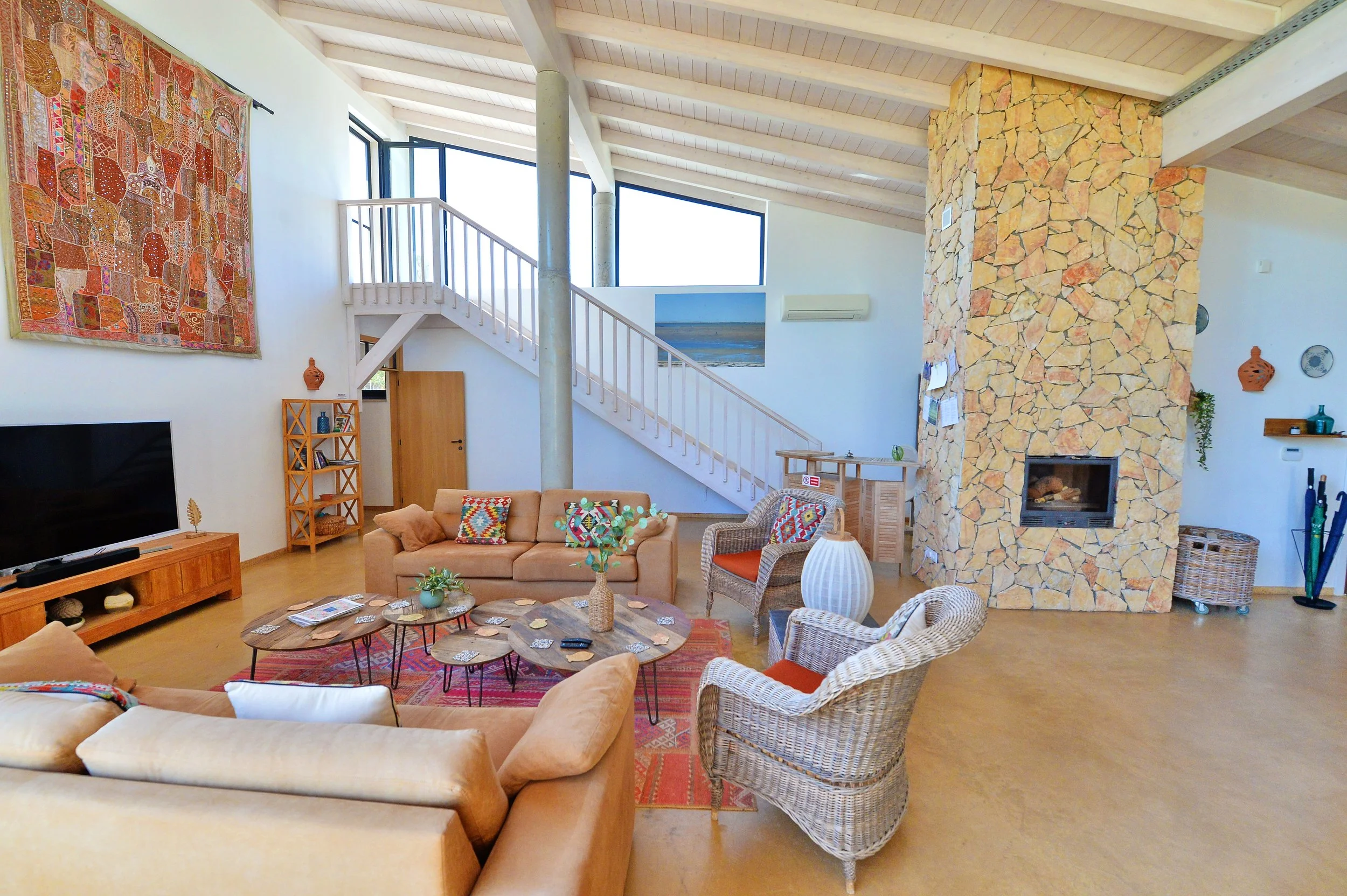 A spacious living room with high vaulted ceiling and large windows. Features a stone fireplace, wicker and upholstered seating, a wood coffee table, a TV on a wooden stand, and colorful decorative pillows and artwork.