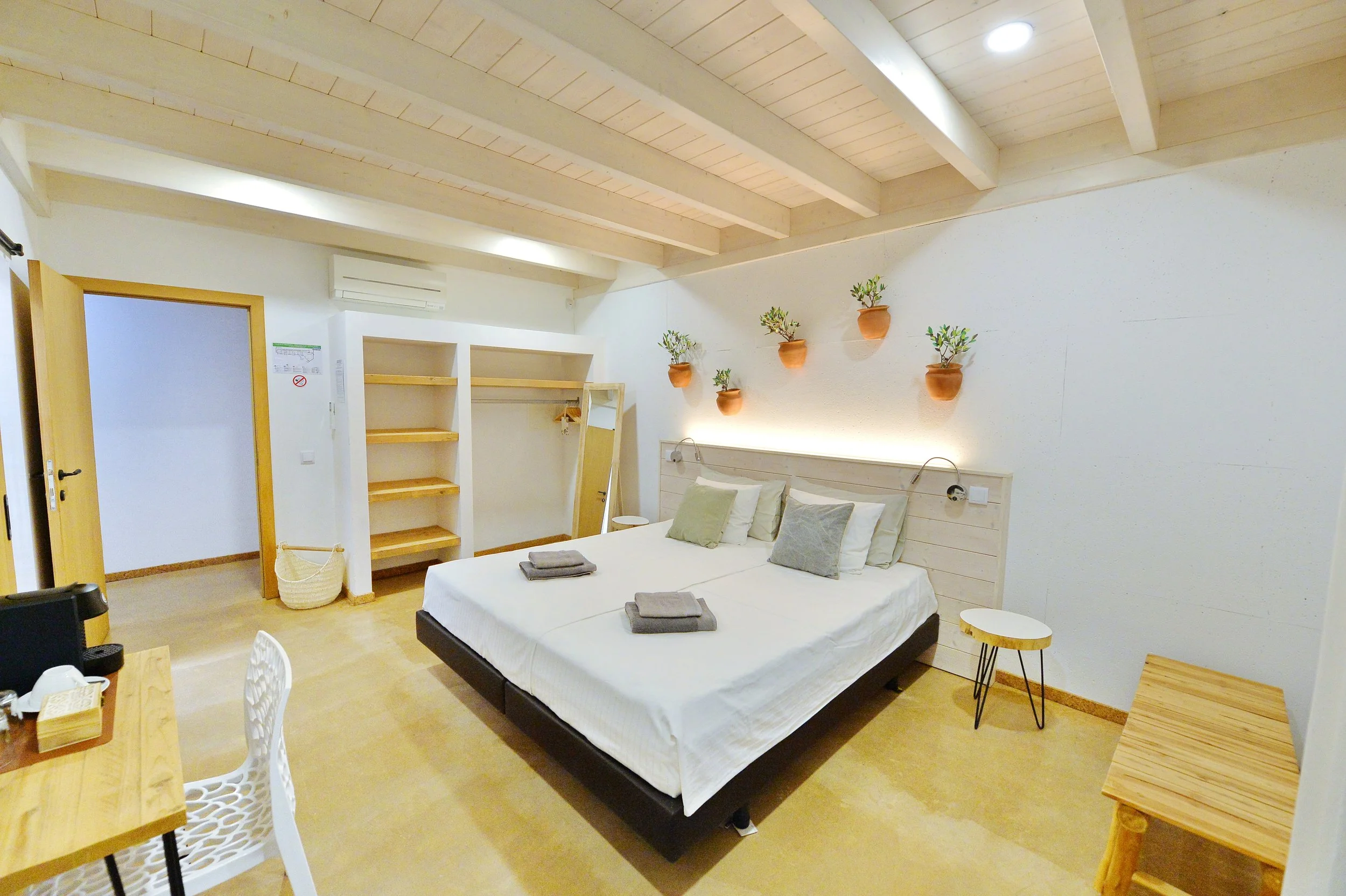 A cozy bedroom with a large bed, white bedding, and multiple pillows. Above the bed are five terracotta wall planters with green plants. The ceiling has exposed wooden beams, and there are shelves and a mirror in the corner. The room features warm wooden furniture and a soft, neutral color palette.