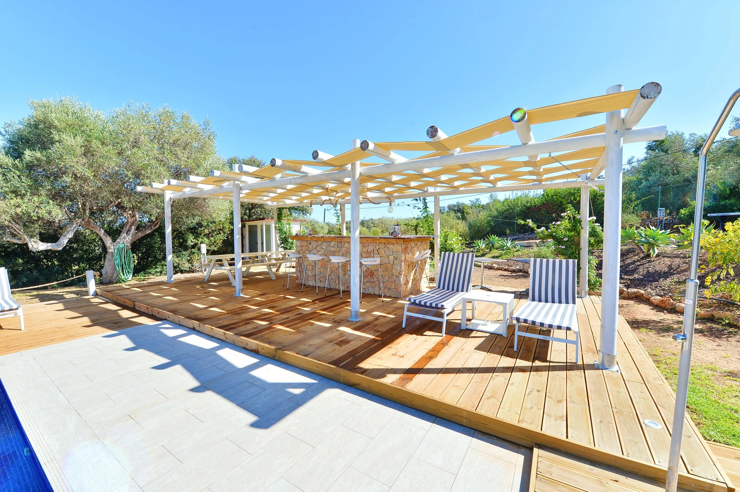 Outdoor patio with wooden deck, lounge chairs with striped cushions, a stone bar area with barstools, and a pergola framework with lights, surrounded by greenery and desert plants.