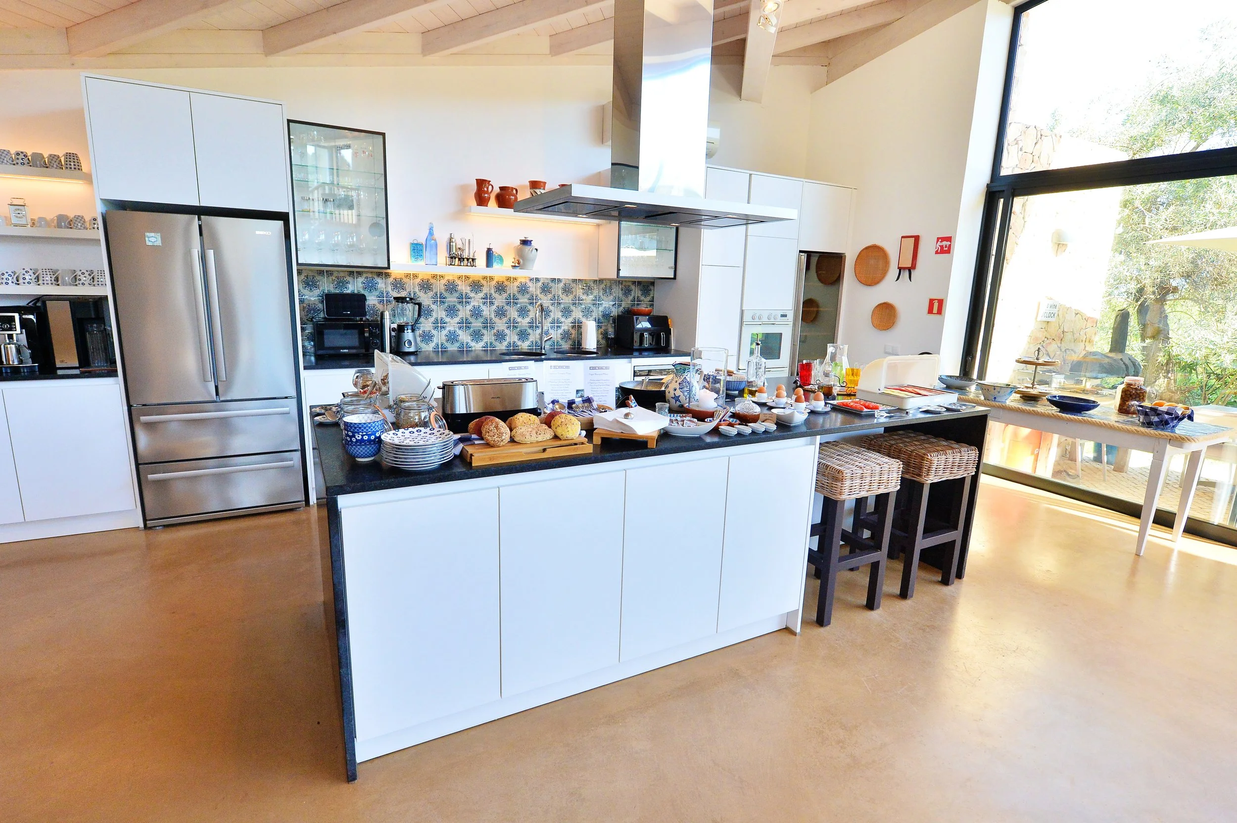 Kitchen with white cabinets, a black countertop, and a stainless steel refrigerator. Countertop has baked goods, dishes, and kitchen appliances. Large window with a view outside.