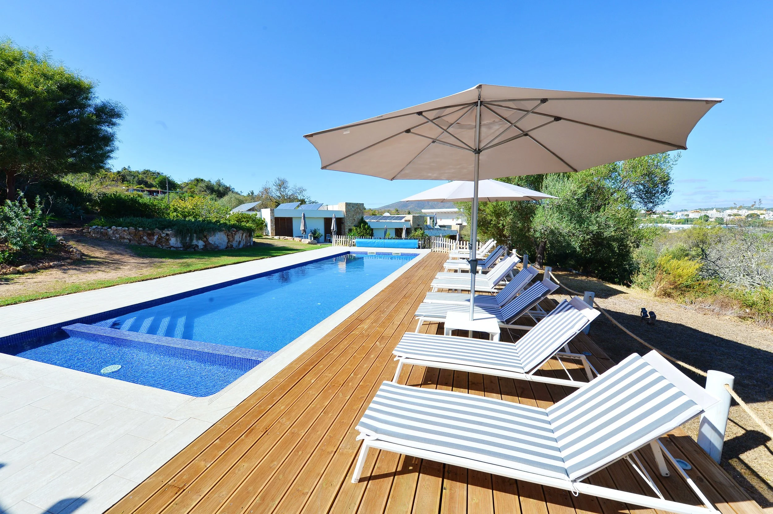 Poolside scene with striped lounge chairs and umbrellas on a wooden deck, overlooking a swimming pool surrounded by greenery under a clear blue sky.