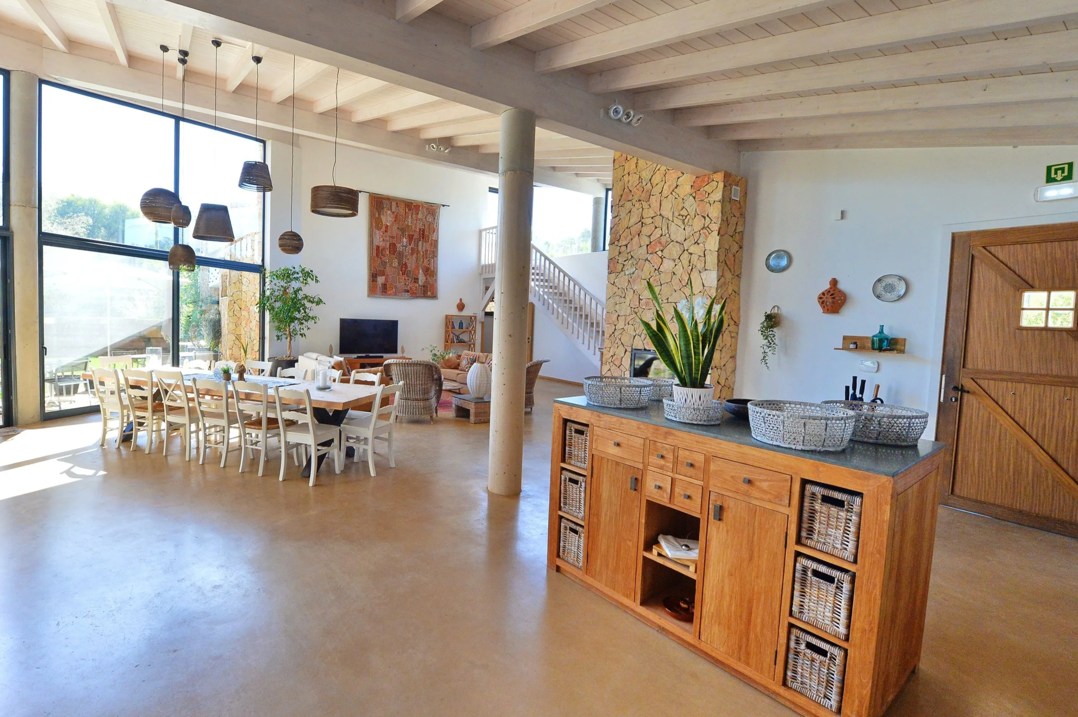 Bright, spacious living and dining area with large windows, wooden beams, and decor featuring woven baskets, potted plants, and wall art.