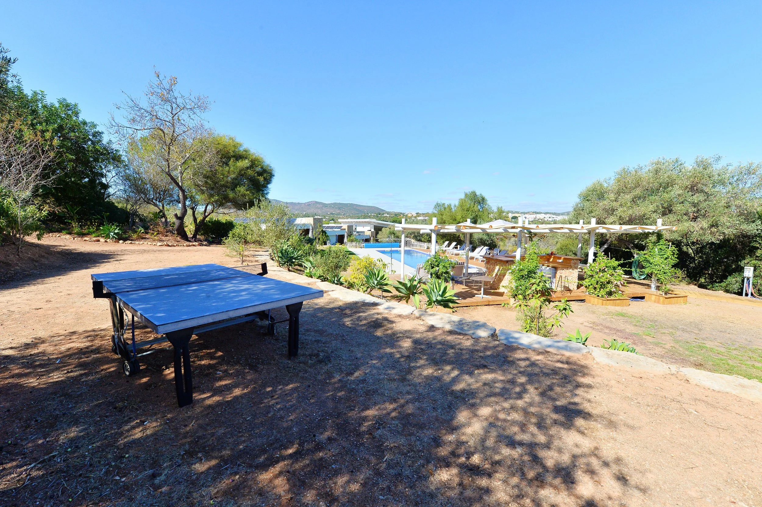 An outdoor scene with a ping pong table on dirt ground in the foreground and a sunlit pool area with lounge chairs, umbrellas, and potted plants in the background, with trees and hills in the distance under a clear blue sky.