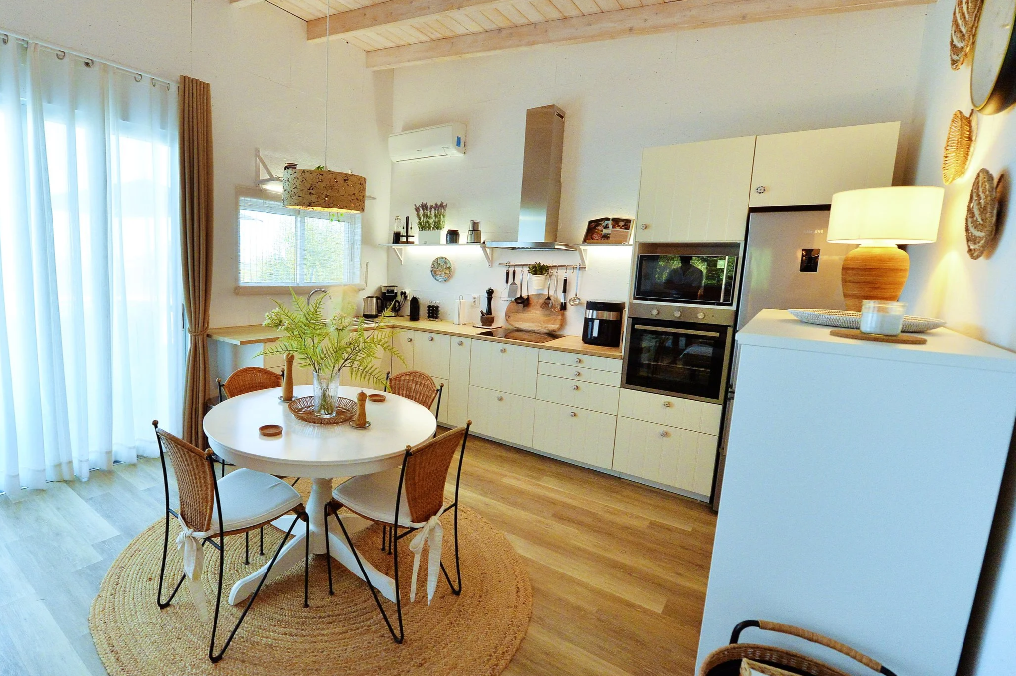 Cozy kitchen with white cabinets, a round dining table with four chairs, a vase with green plants, and a sliding glass door with curtains.