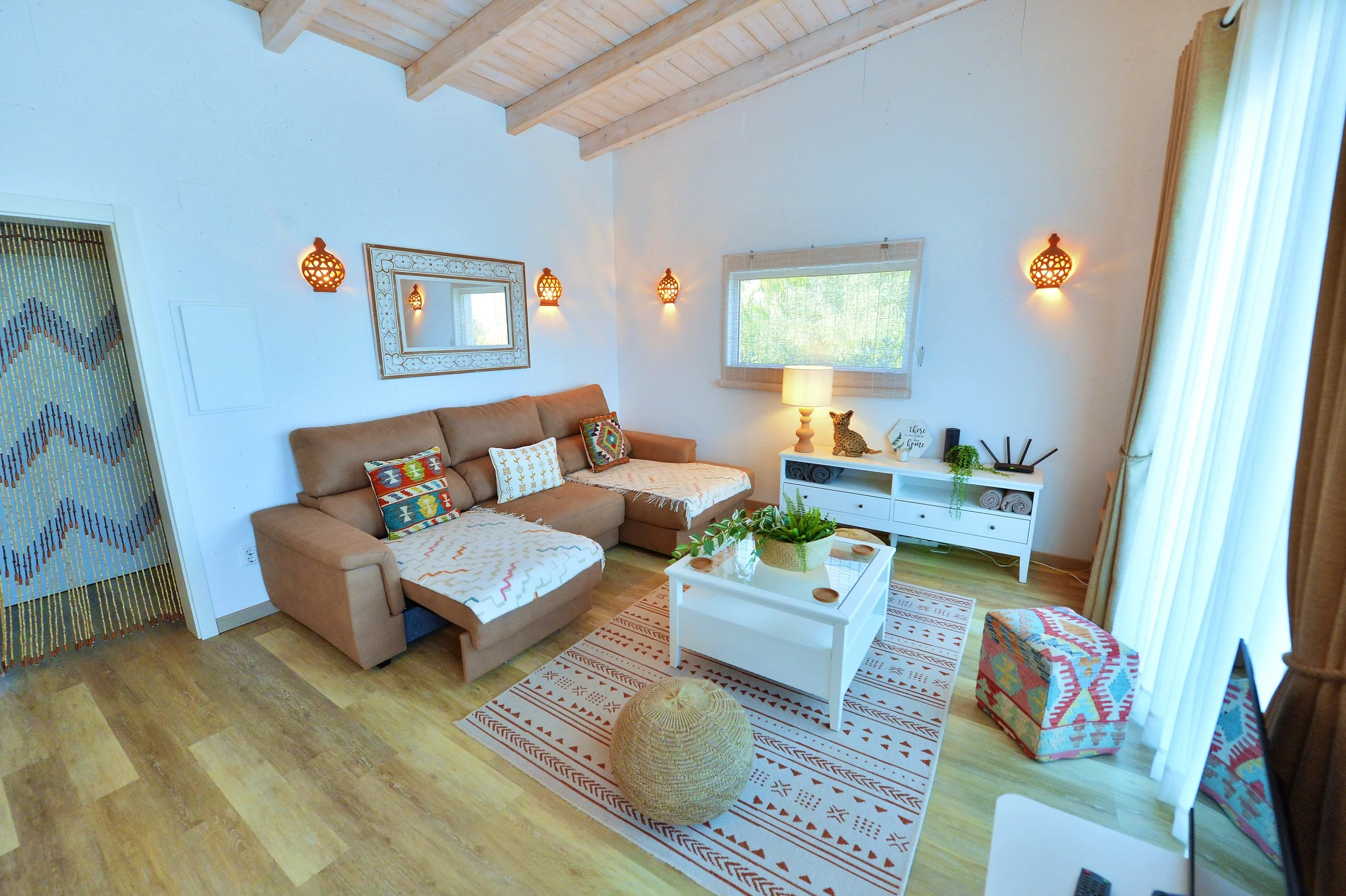 Living room with a tan sectional sofa, patterned pillows, wooden ceiling beams, white coffee table with green plants, white sideboard with lamp and decorative items, rug with geometric pattern, curtain-covered windows, and a beaded curtain partition.
