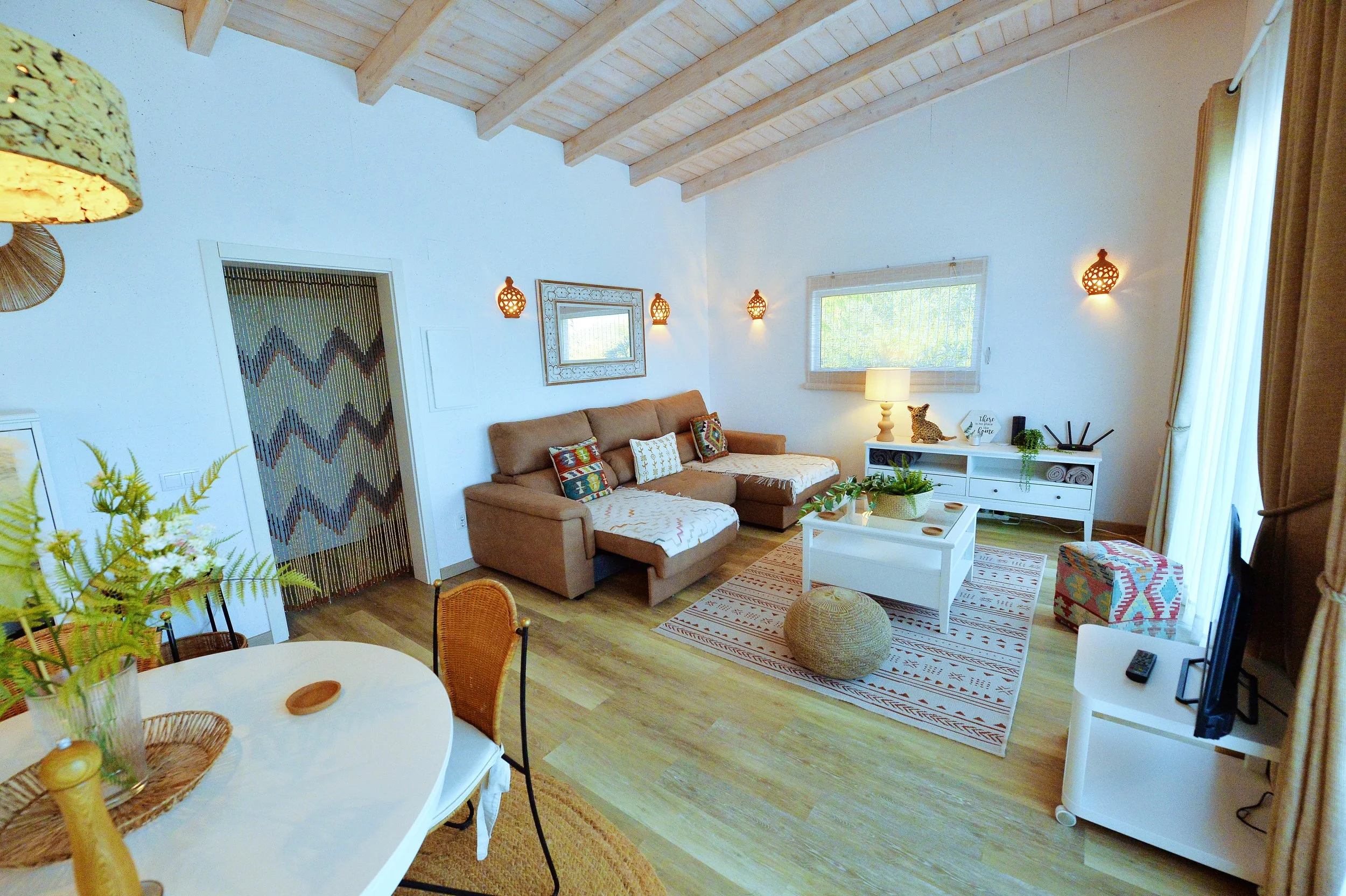 Living room with beige sectional sofa, white coffee table, patterned throw pillows, and a white sideboard with decorative items. Wooden ceiling with exposed beams, light-colored floor, window with blinds, and a sliding glass door with curtains. Warm 