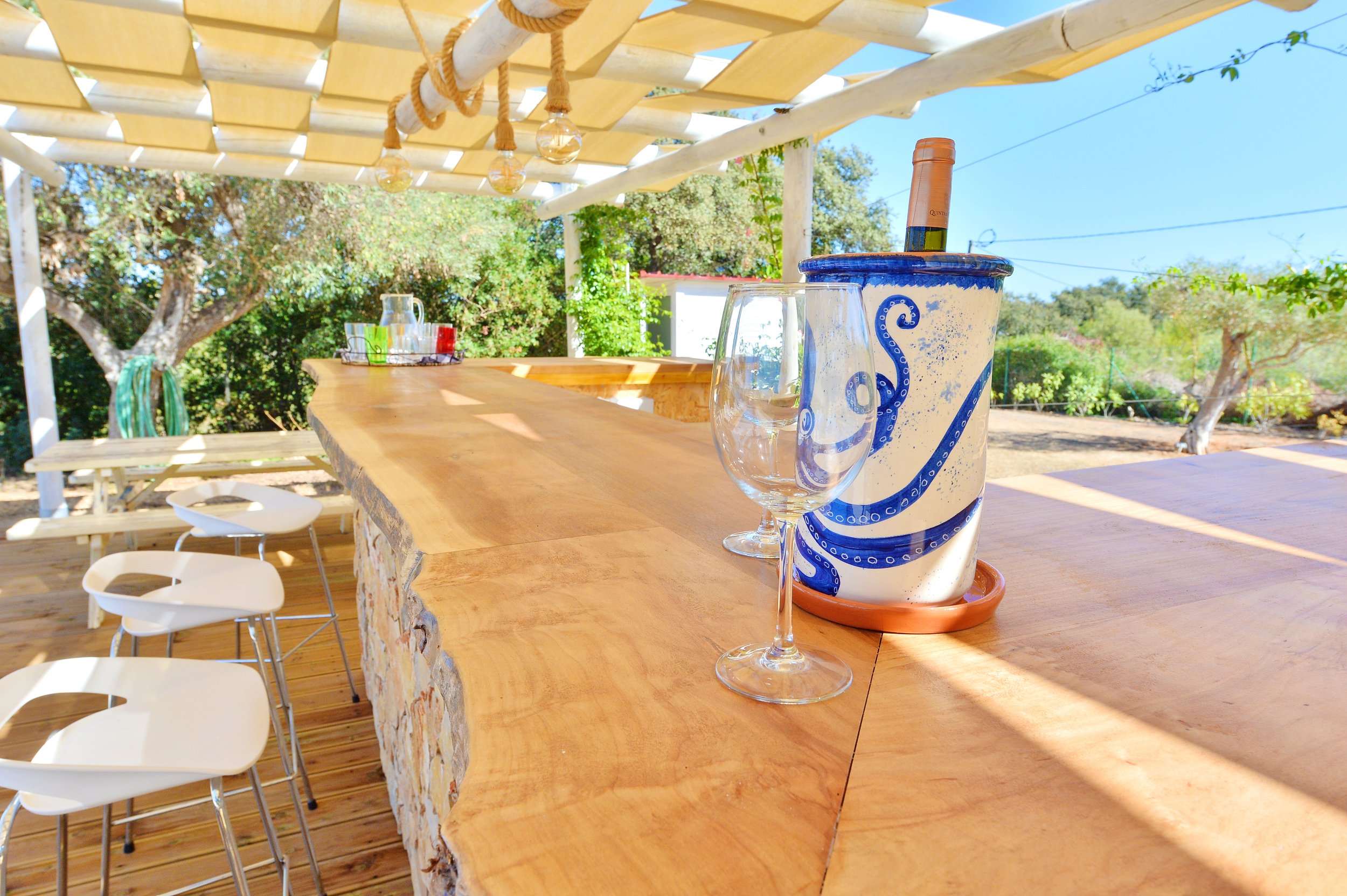 Outdoor bar area with a wooden bar counter, a wine bottle in an ice bucket, an empty wine glass, and a glass pitcher with drink mixers. There are bar stools lined up in front, and a shaded overhead with string lights. Trees and a clear blue sky are visible in the background.