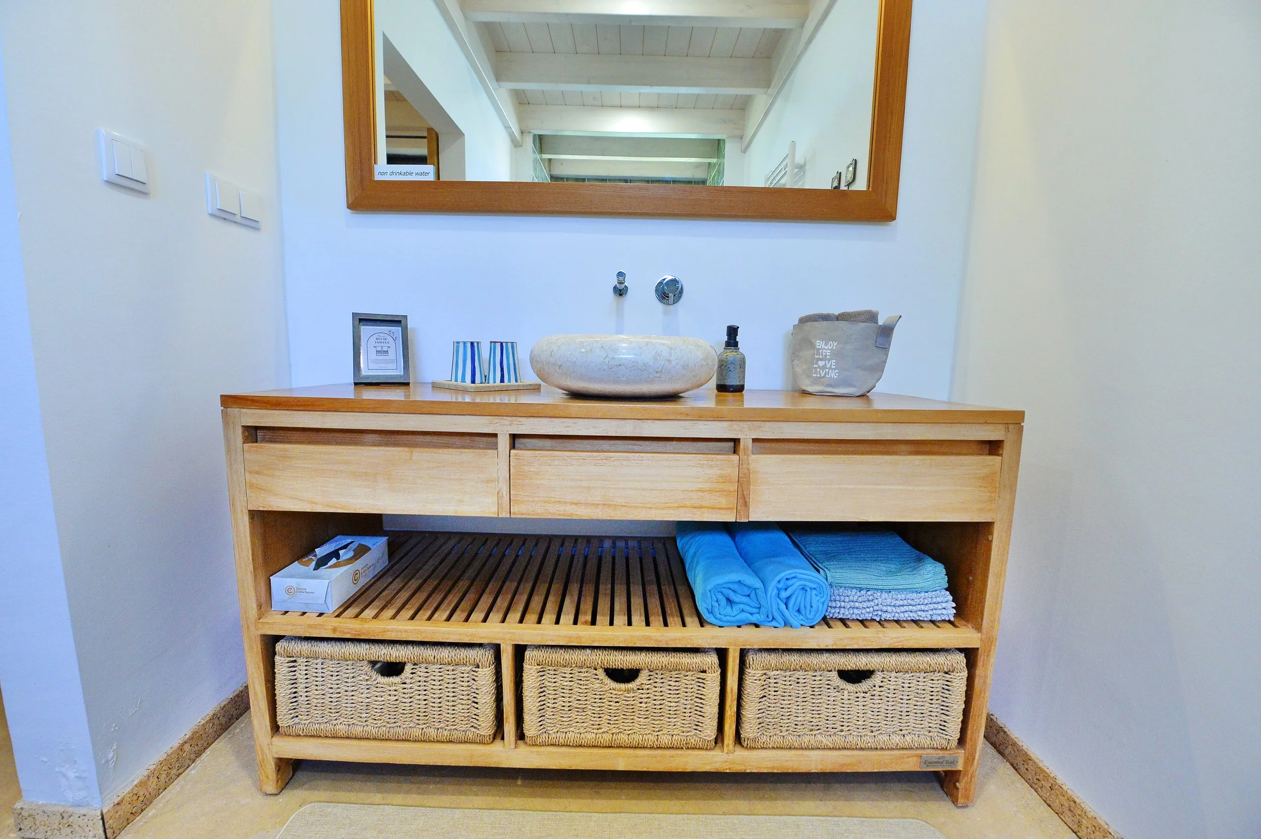 Wooden bathroom vanity with a round stone sink, soap dispenser, and decoration items parked on top. A large mirror hangs above, reflecting ceiling beams and hallway. Lower shelves contain rolled towels and wicker baskets.