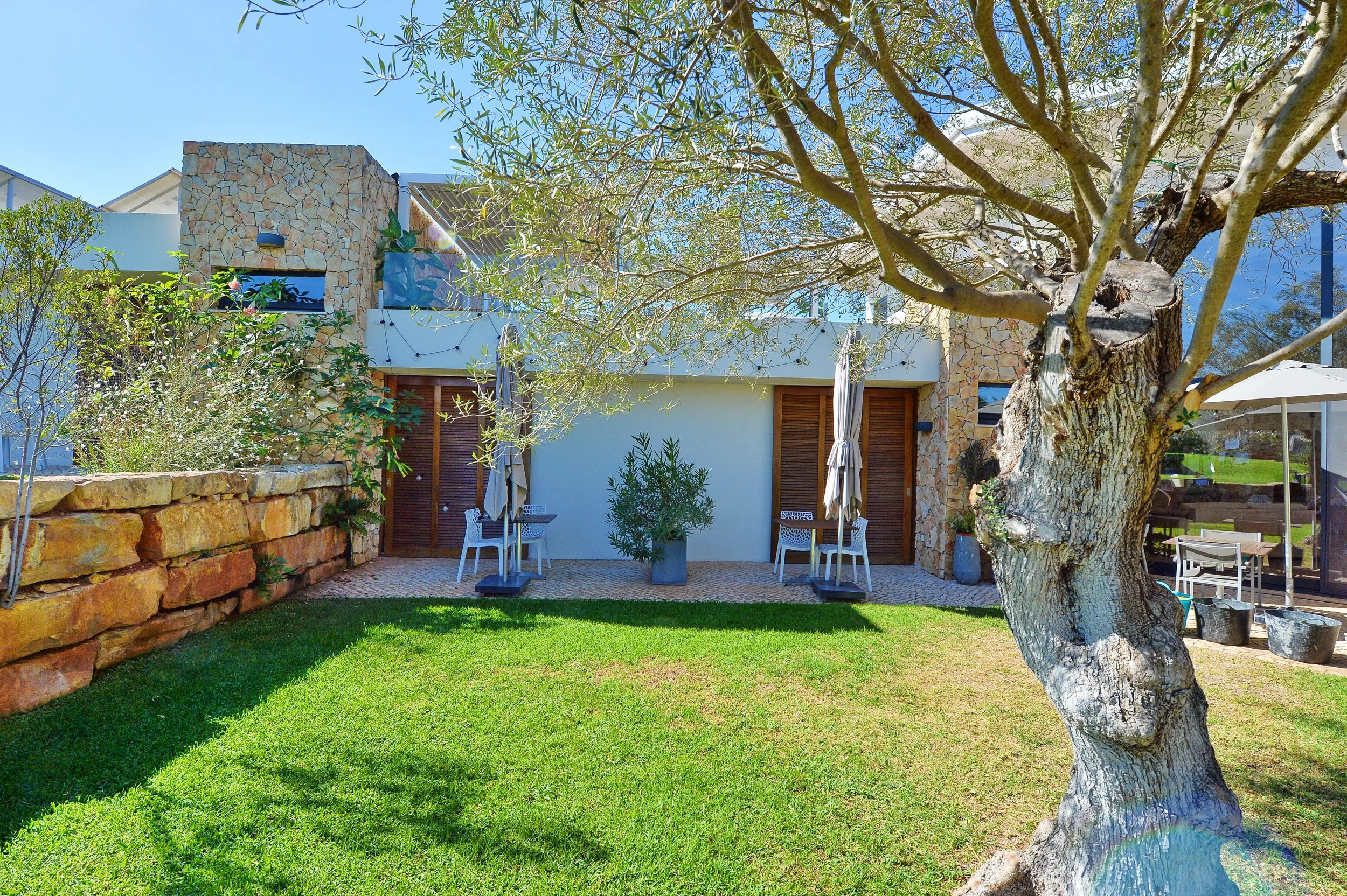 A backyard garden with a large, moss-covered tree in the foreground, green grass, a small patio with two white chairs and umbrellas, a potted plant in the middle, a stone wall on the left, and a house with windows, a balcony, and a glass door on the 