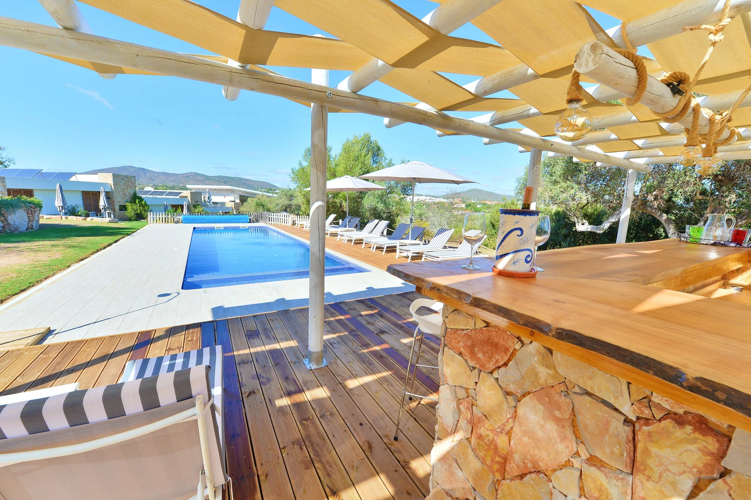 Outdoor pool area with lounge chairs, umbrellas, and a bar with a wooden countertop. Wooden and stone flooring with a decorative pergola overhead. Green trees and distant mountains in the background on a sunny day.