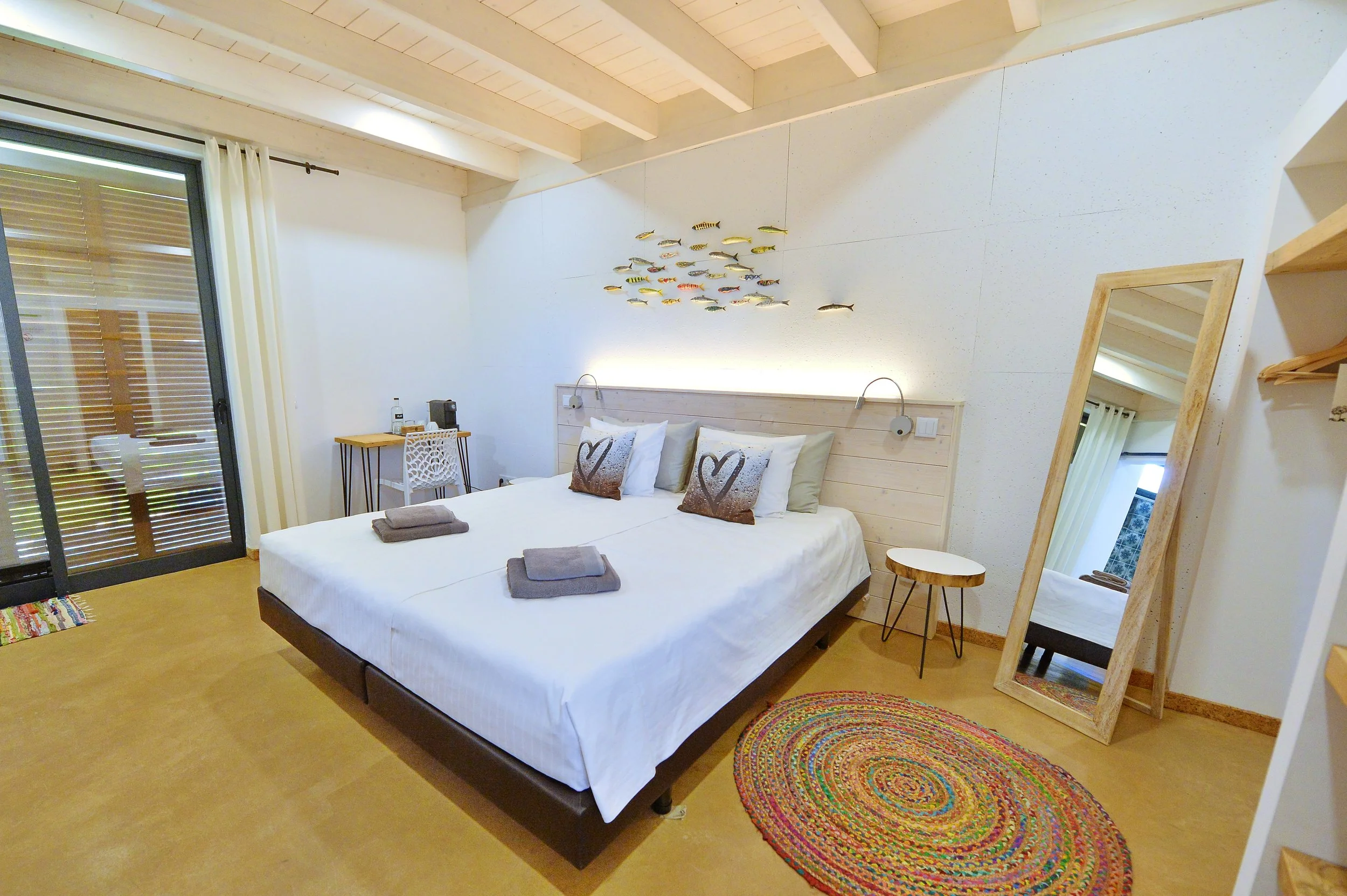 A cozy bedroom with a large bed, two pillows with heart designs, and towels on top. There is a wooden headboard, a mirror leaning against the wall, a small side table, a colorful oval rug, and a fish wall decoration. The room has a sliding glass door with curtains and a small desk in the corner.