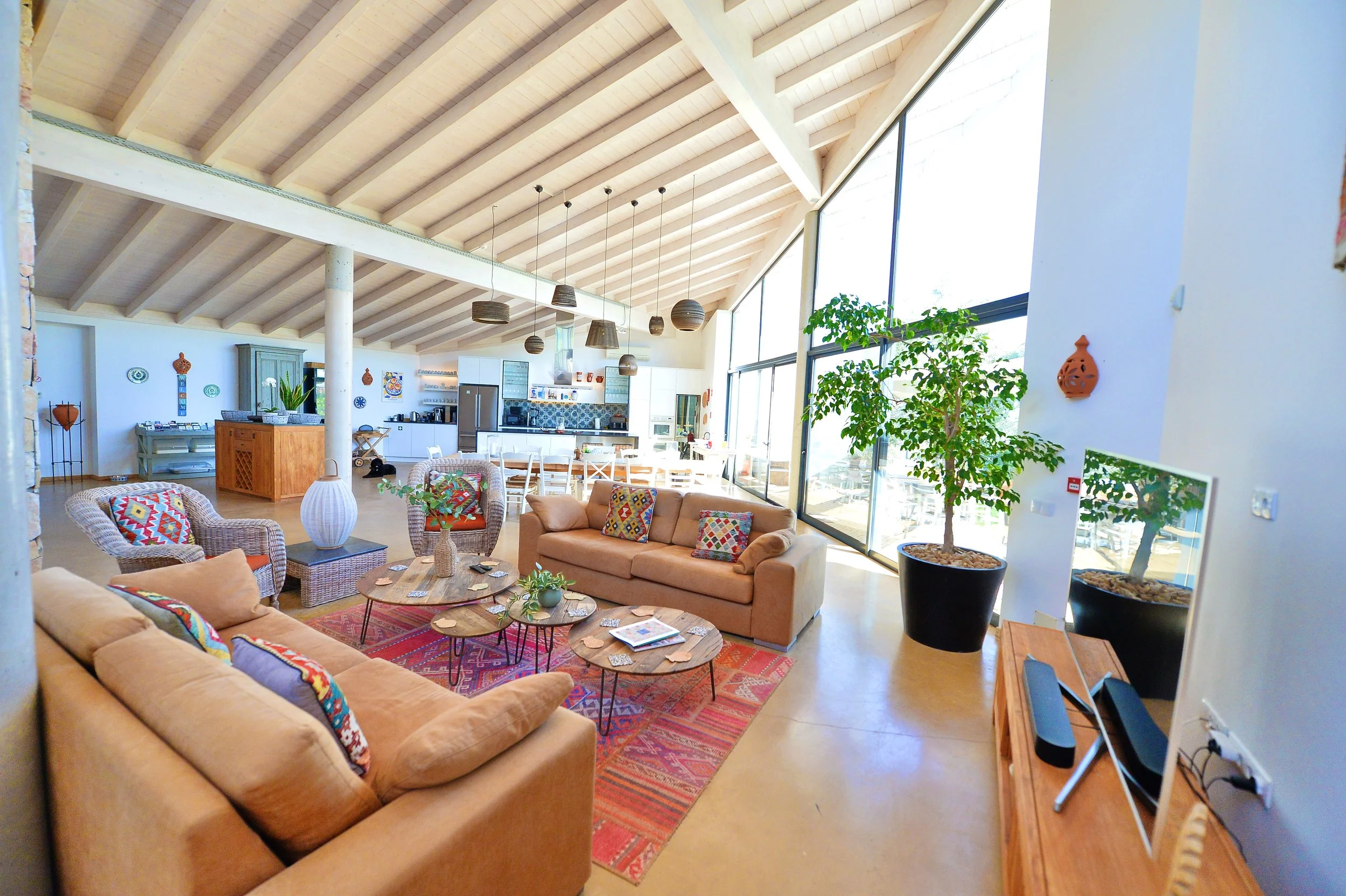 Bright open living room with large windows, beige sofas with colorful pillows, a red patterned rug, coffee tables, and a kitchen in the background with white cabinets and a wooden island.