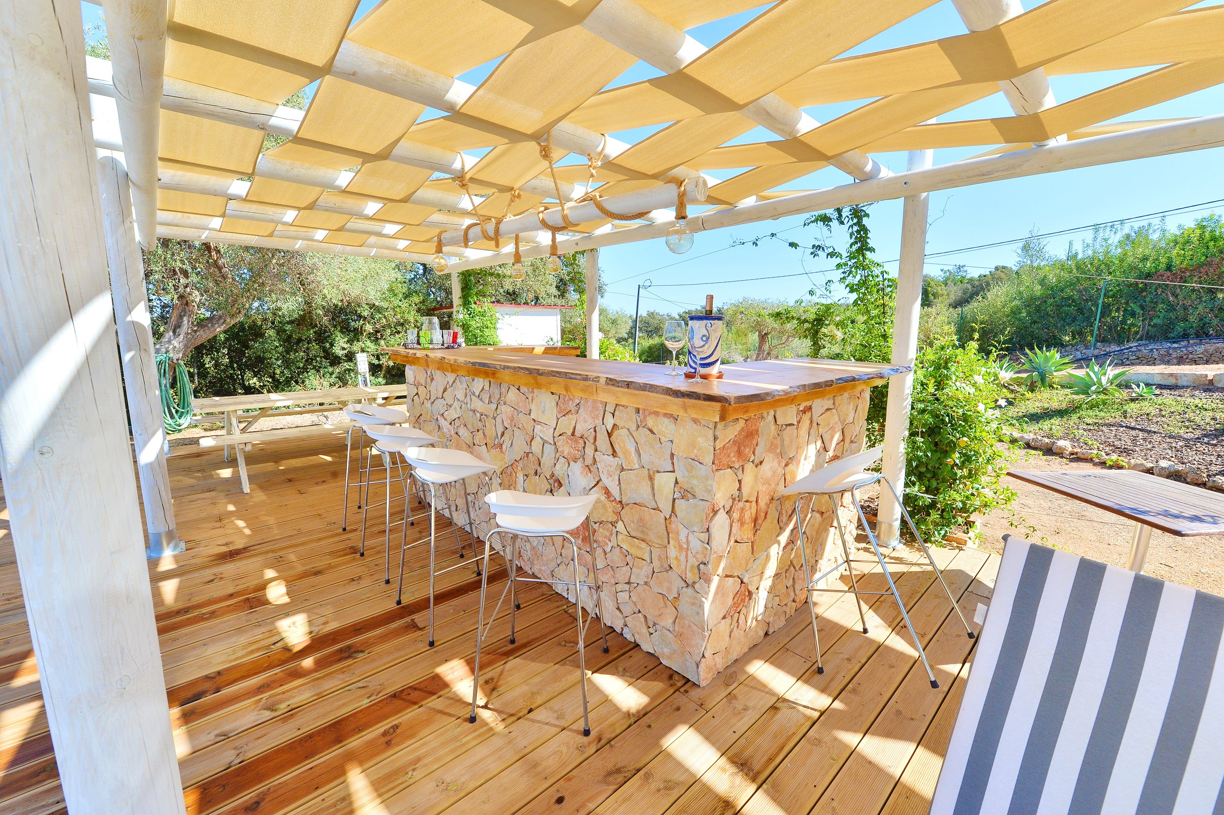 Outdoor patio area with a stone bar, white bar stools, a wooden pergola with hanging bulbs, surrounded by greenery and desert plants.