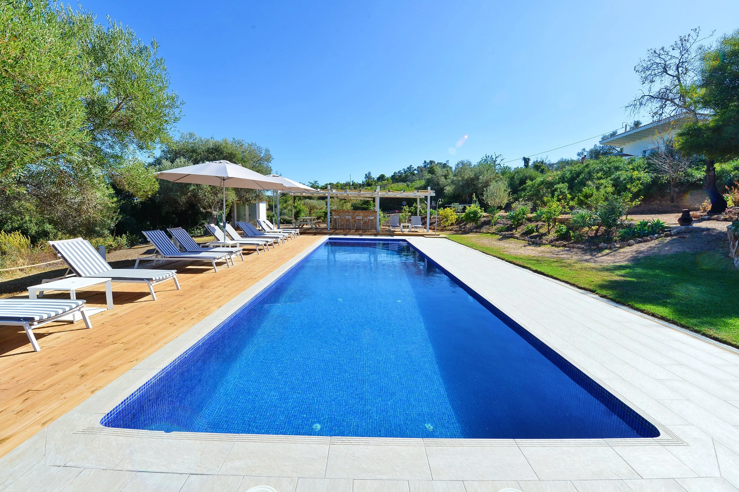 Outdoor swimming pool with a wooden deck, surrounded by lounge chairs with striped cushions, an umbrella, and lush greenery on a sunny day.