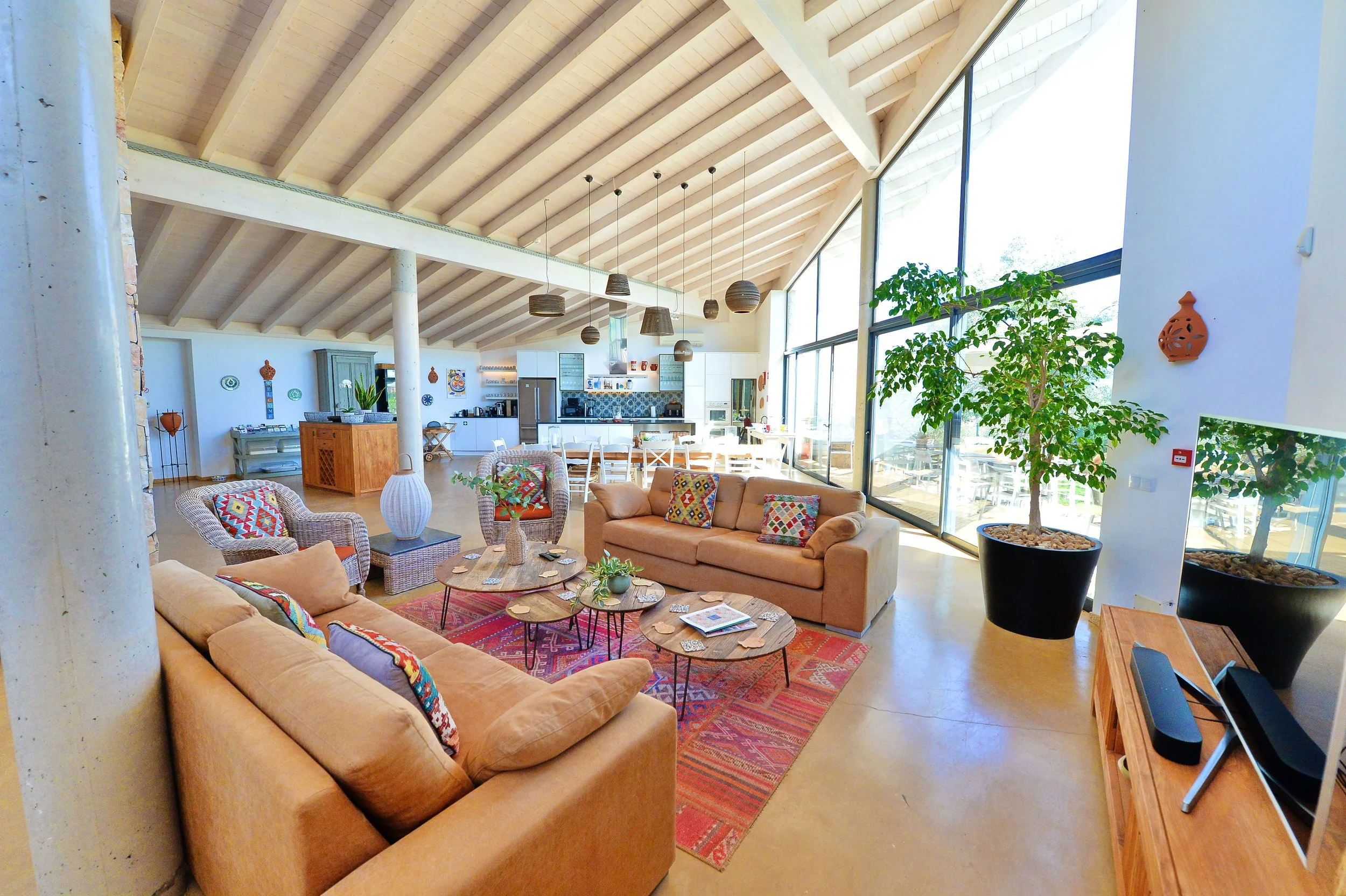 A spacious open-concept living area with high, vaulted wooden ceilings, large glass windows, and modern furniture including beige sofas and patterned armchairs. There is a large potted plant next to the windows, and a kitchen with white cabinetry and a dining table in the background. Decor includes colorful pillows, a patterned rug, and hanging pendant lights.