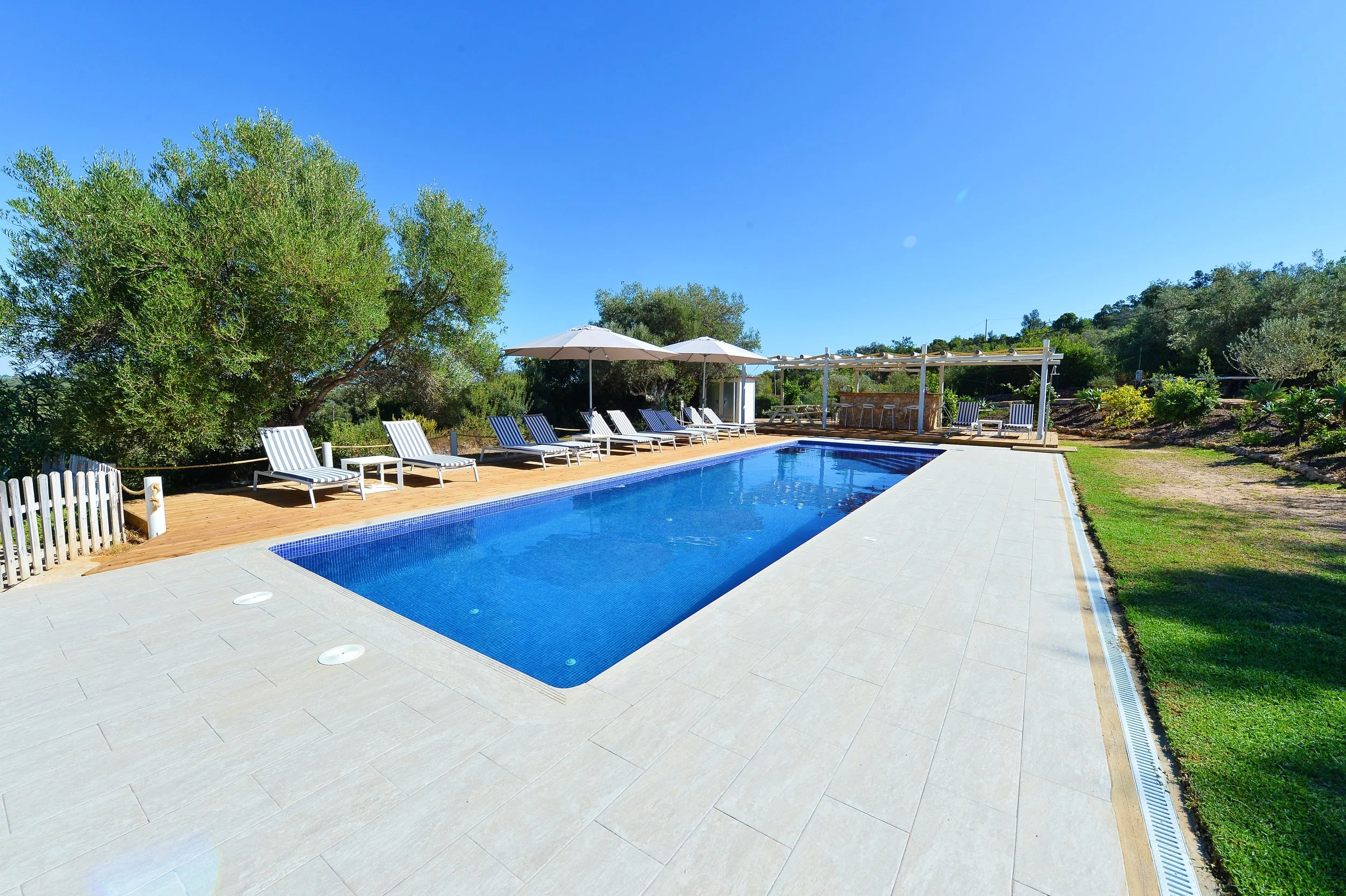 Outdoor swimming pool surrounded by lounge chairs and umbrellas, with greenery and trees in the background under a clear blue sky.