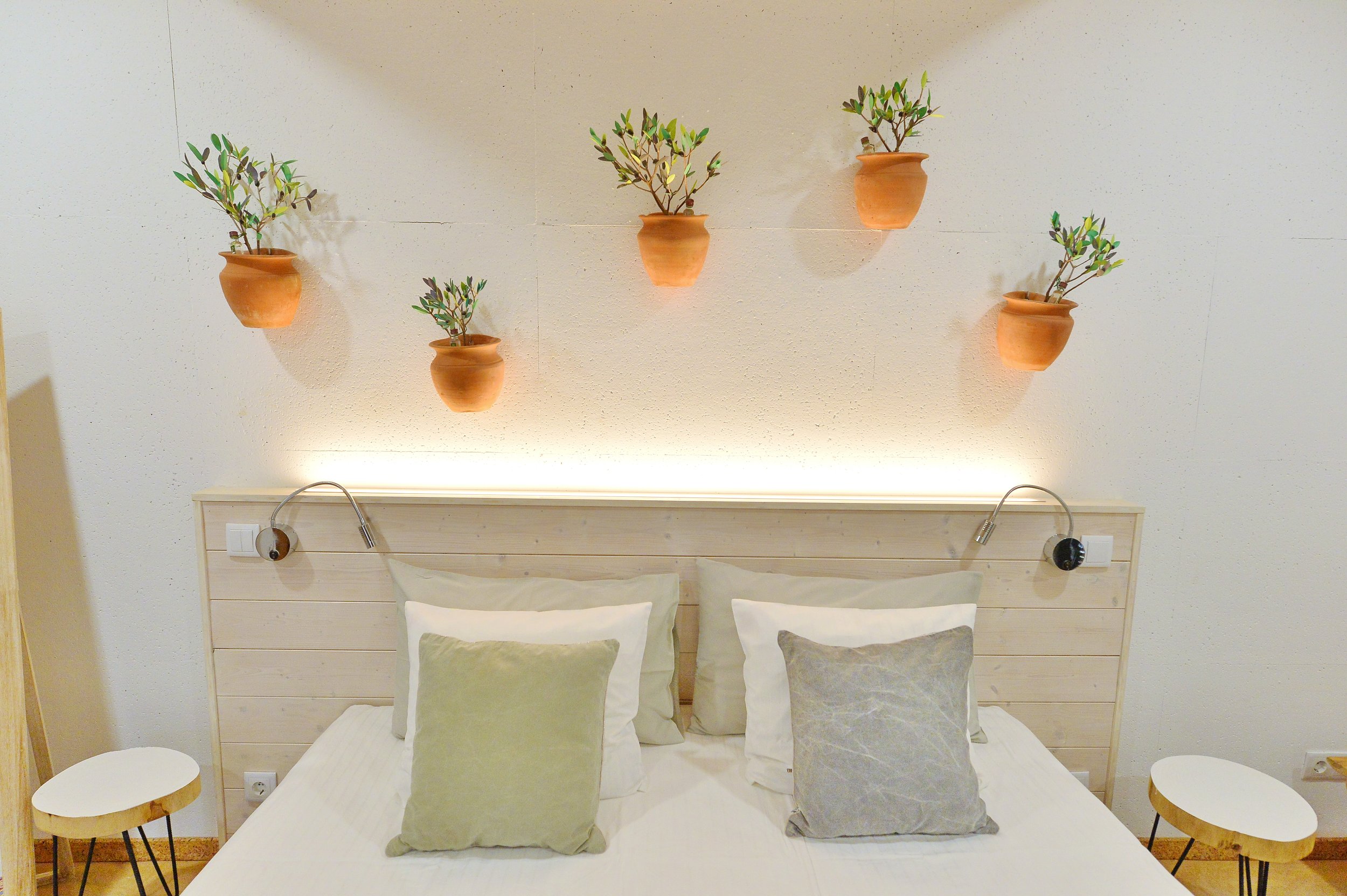 A modern bedroom with a light wooden headboard, four beds, and two bedside tables. Above the bed, five small terracotta pots with green plants are mounted on the wall, and soft lighting illuminates the space.