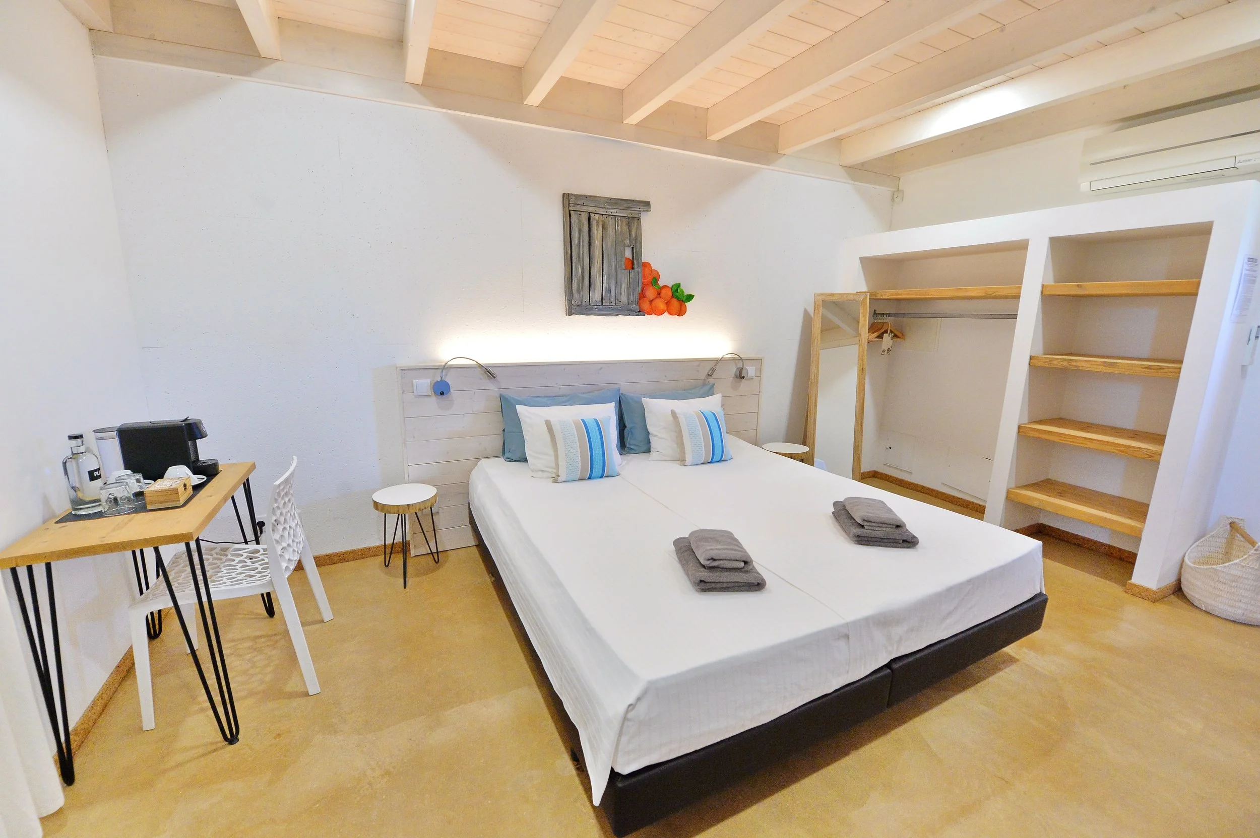 Hotel room with a double bed, white bedding, gray and blue pillows, two folded gray towels, a small wooden desk with a coffee machine, water bottles, and glasses, a white chair, a small round side table, and a wooden shelving unit in the corner.