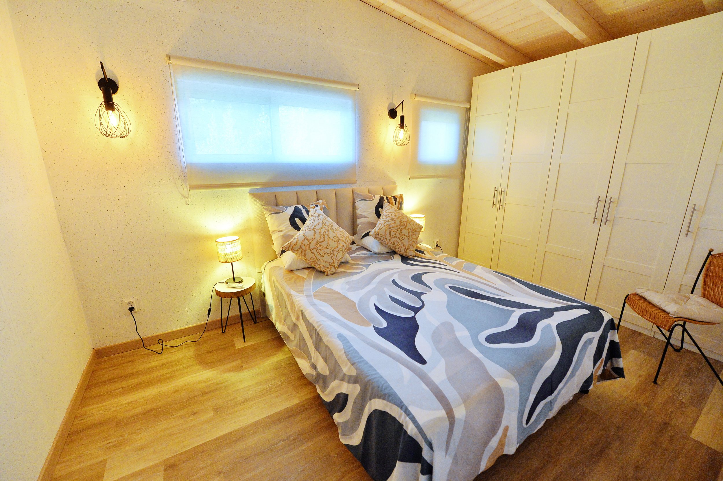A cozy bedroom with a bed that has beige and patterned pillows, a blanket with abstract patterns, and two small bedside lamps. The room features wood flooring, two wall-mounted lights, a large built-in wardrobe, a small wicker chair, and three small windows with blinds and curtains.