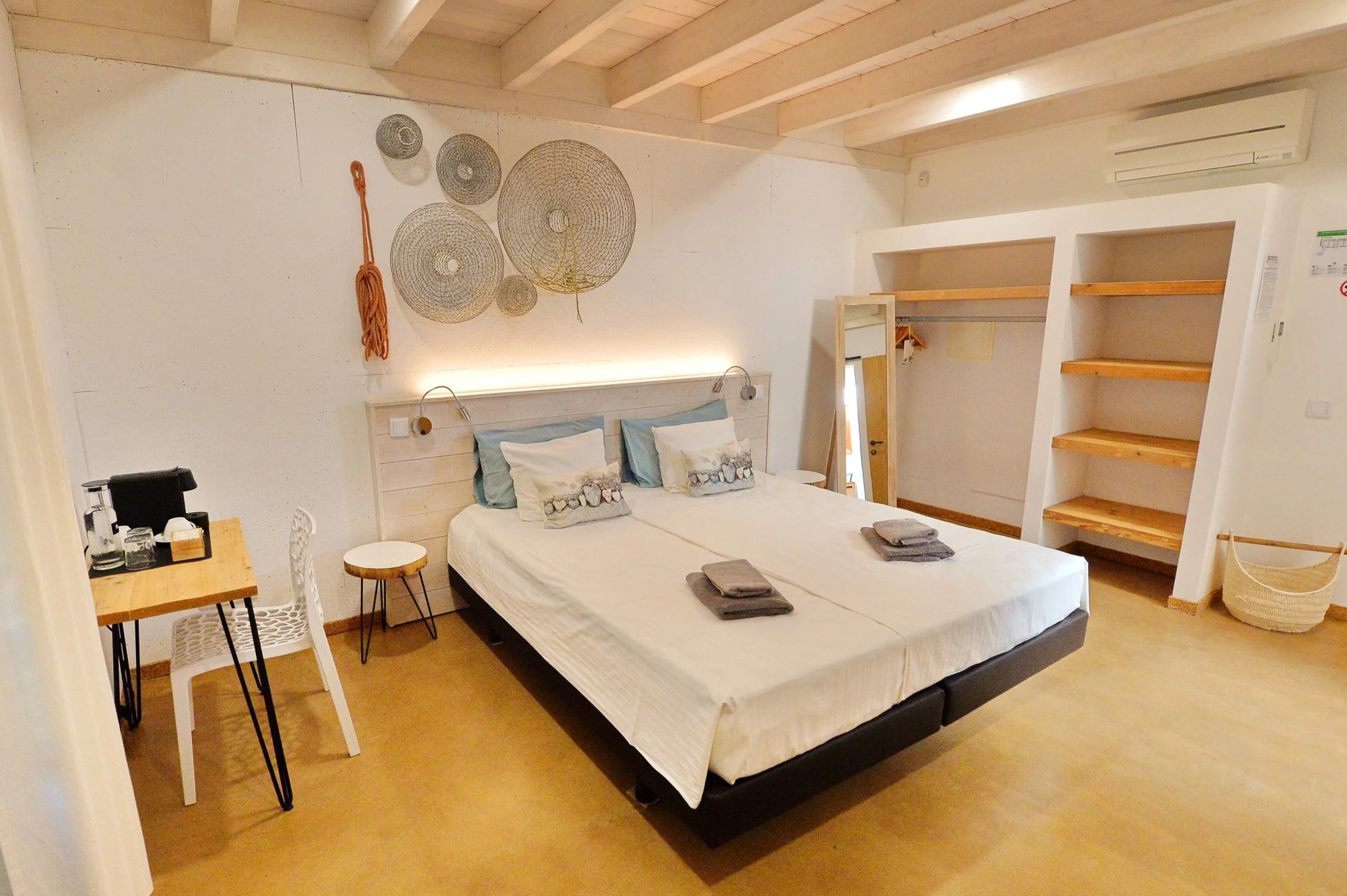 A bedroom with a double bed, white bedding, pillows, and folded towels on the bed. The room has wooden shelves, a mirror, and decorative wall hangings. There is a small table with a chair and a stool, and an air conditioning unit on the wall.
