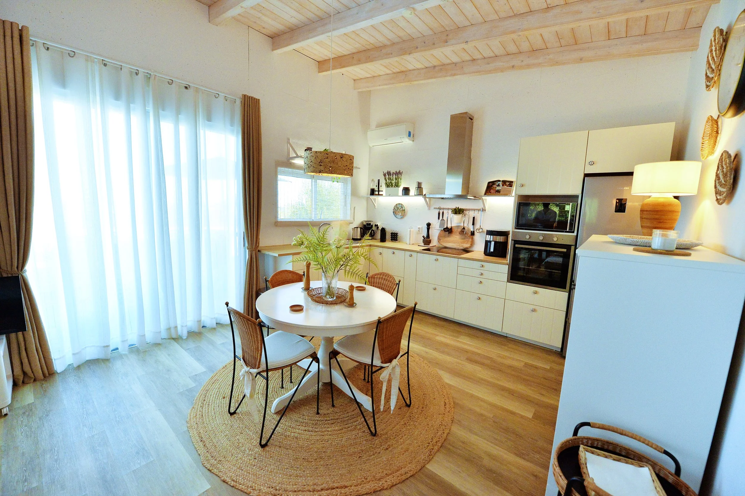 Bright kitchen and dining area with white cabinetry, a round dining table with four chairs, and large windows with sheer white curtains and brown drapes.