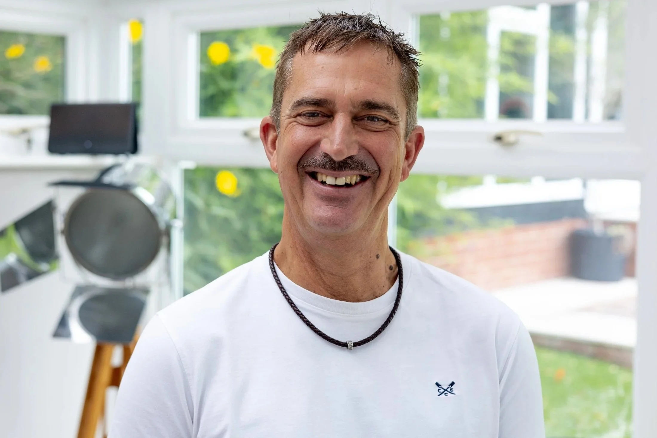 A smiling man with short brown hair, wearing a white t-shirt and a black necklace, standing inside a bright room with large windows and a blurred outdoor background.