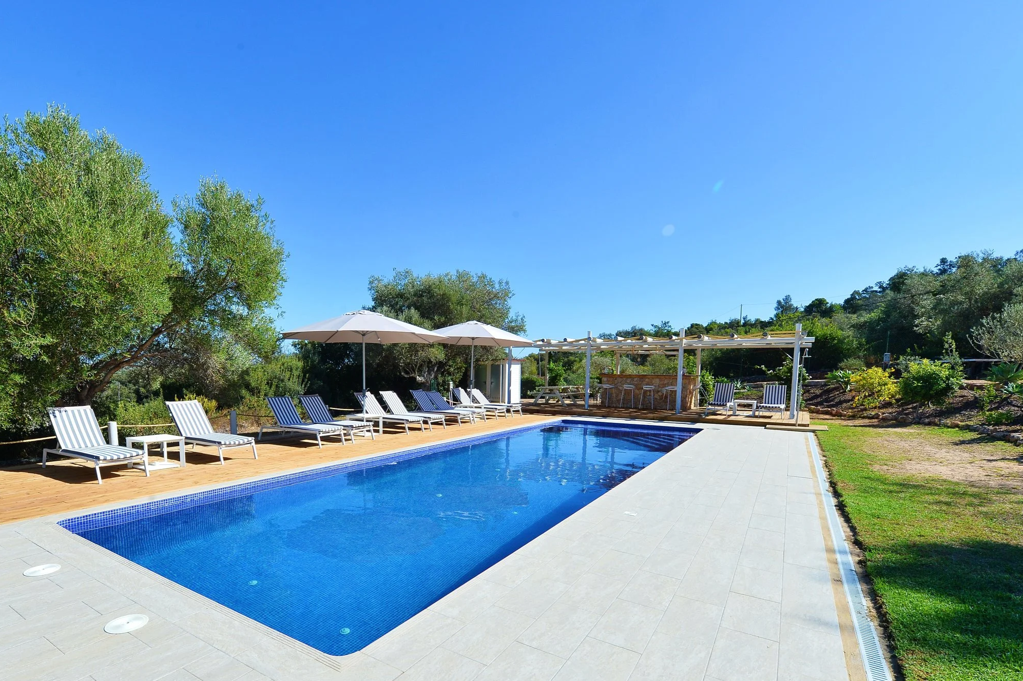 Outdoor swimming pool with blue water, surrounded by lounge chairs with blue and white striped cushions, large white umbrellas, and a shaded bar area with seating, set against green trees and a clear blue sky.