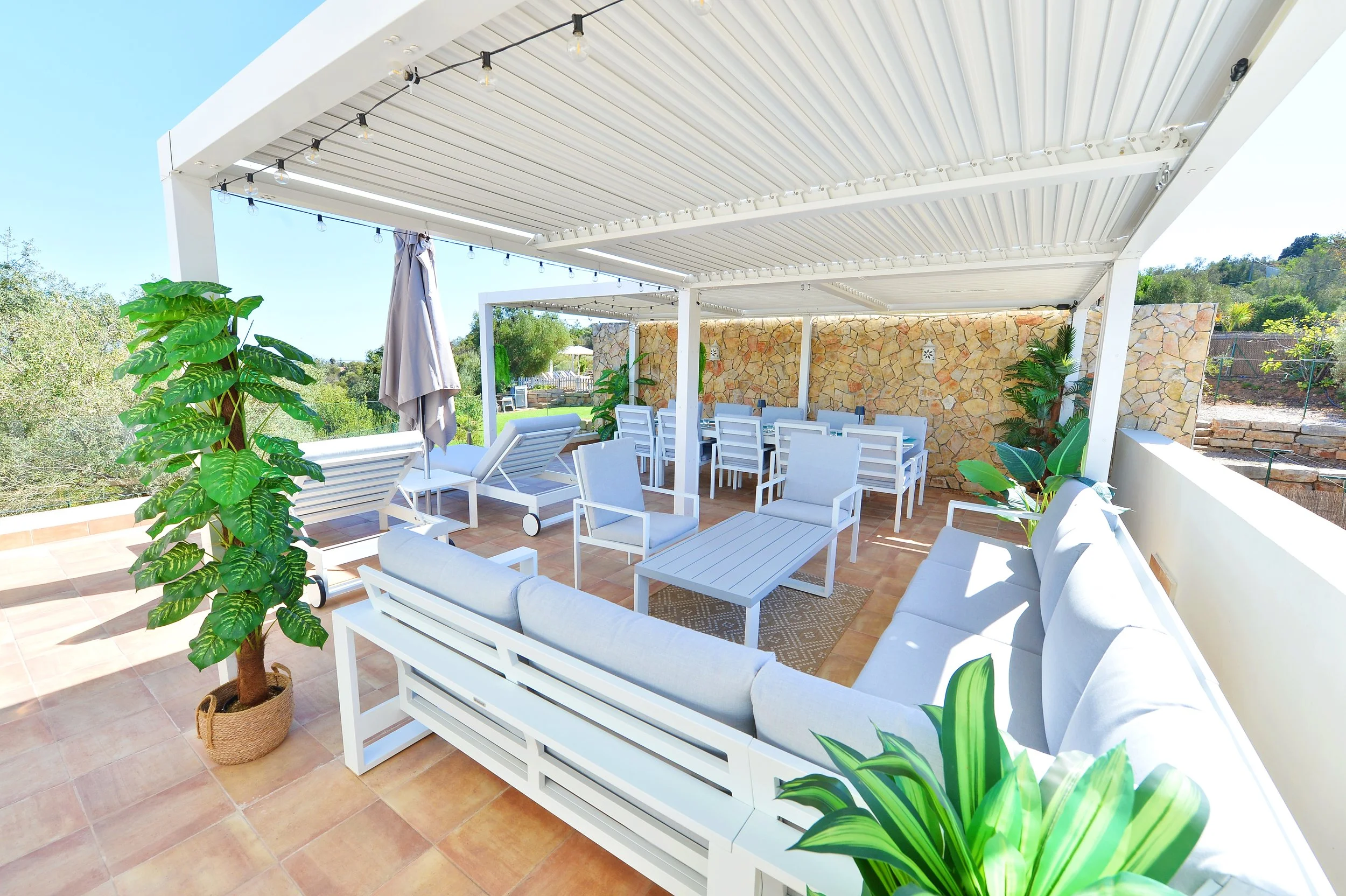 Outdoor patio with white furniture, potted plants, and a stone wall, shaded with a white pergola and string lights.