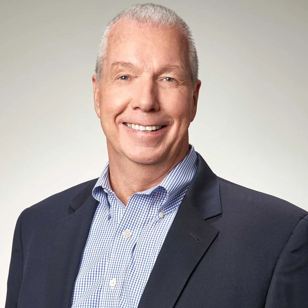 Portrait of a smiling middle-aged man with short gray hair, wearing a dark blazer and a checkered dress shirt, against a light, neutral background.