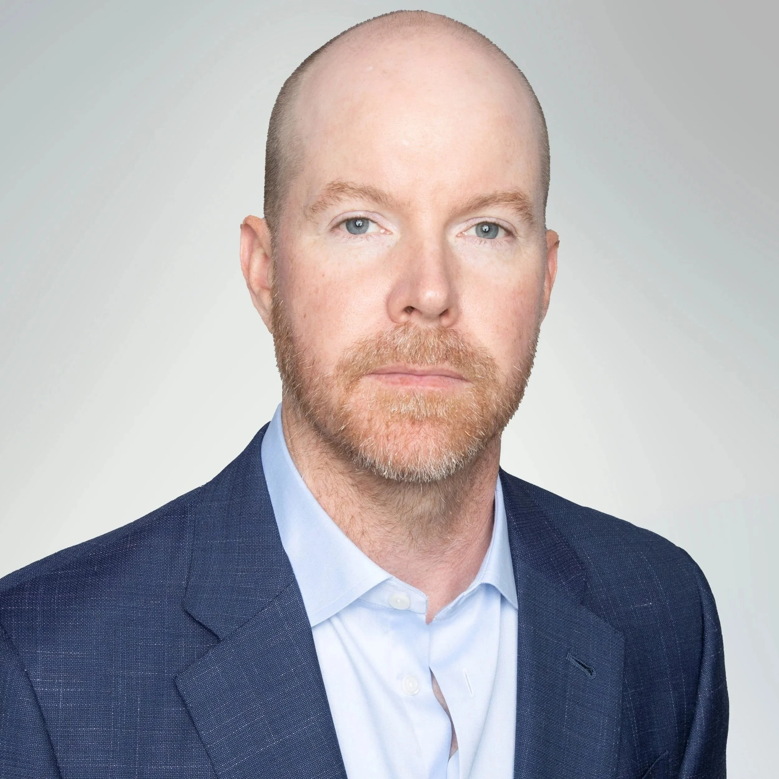 Headshot of a man with red hair and beard wearing a dark blue suit and light blue shirt against a plain light gray background.
