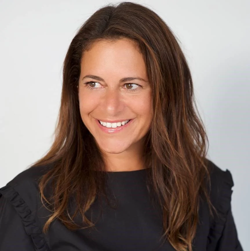 Headshot of a smiling woman with brown hair, wearing a black top, against a neutral background.