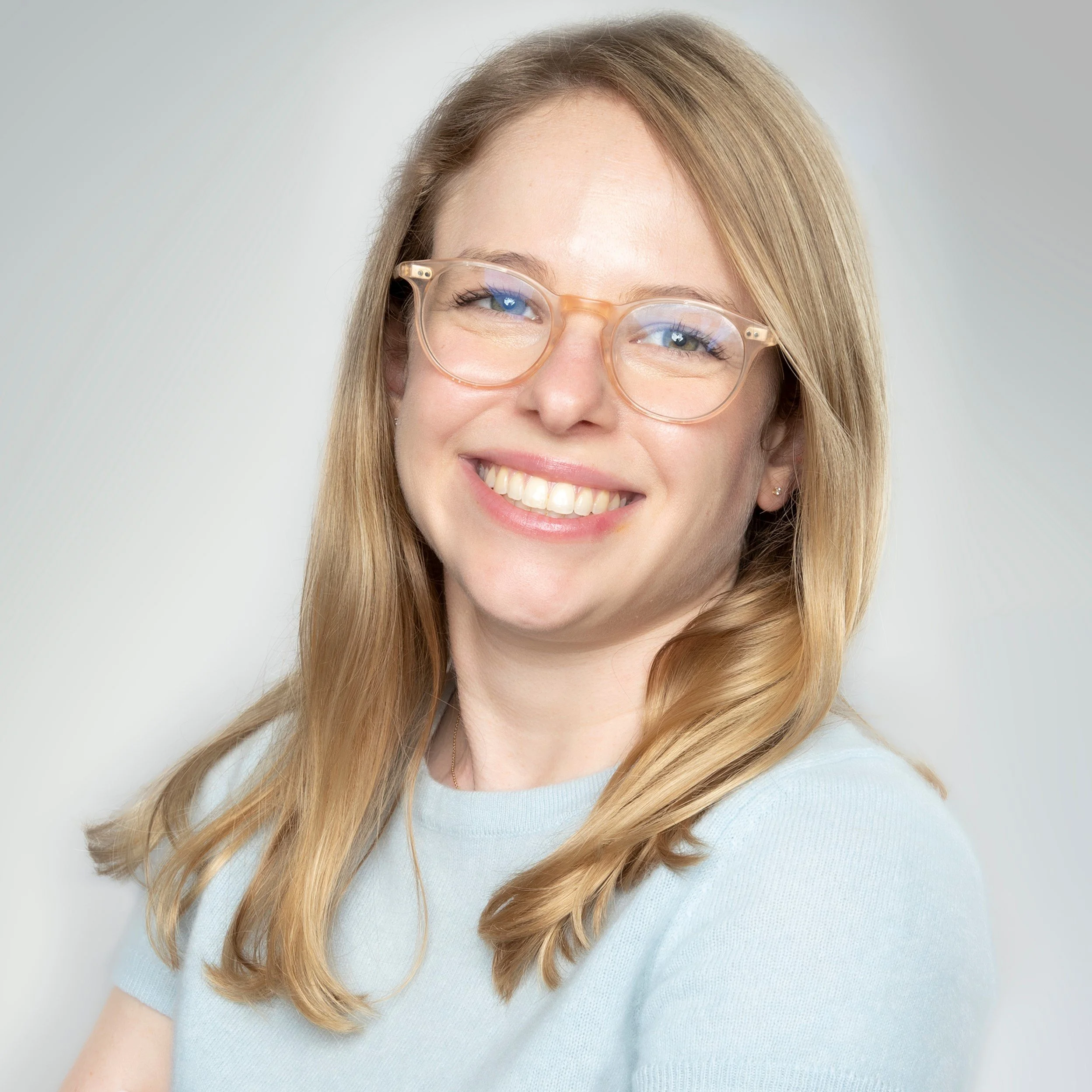 Close-up portrait of a smiling woman with long, wavy blonde hair, wearing clear glasses and a light blue top, against a neutral background.