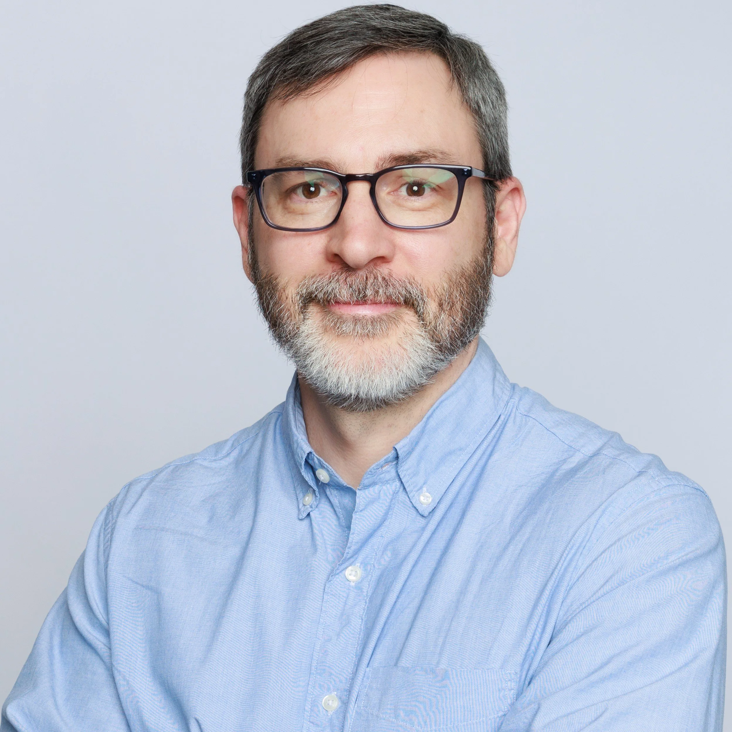 Portrait of a man with glasses, a beard, and gray hair, wearing a light blue button-up shirt, standing against a plain light gray background.