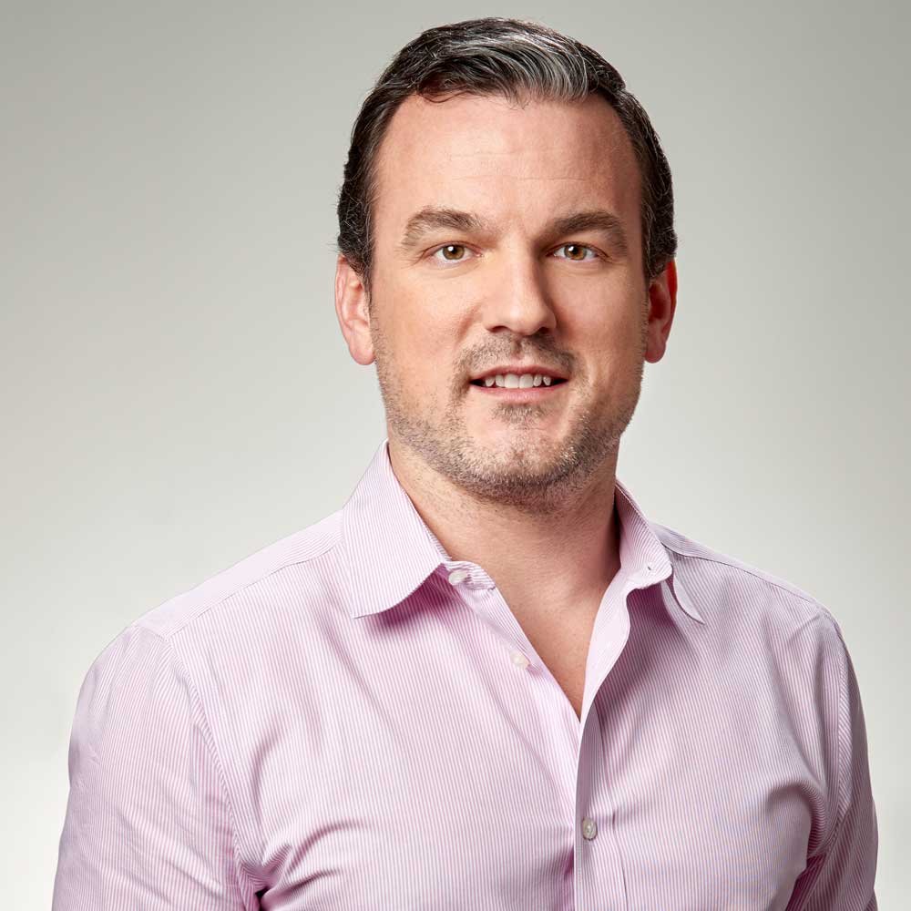 A man with short dark hair, a light beard, and wearing a pink dress shirt, smiling at the camera against a plain light background.