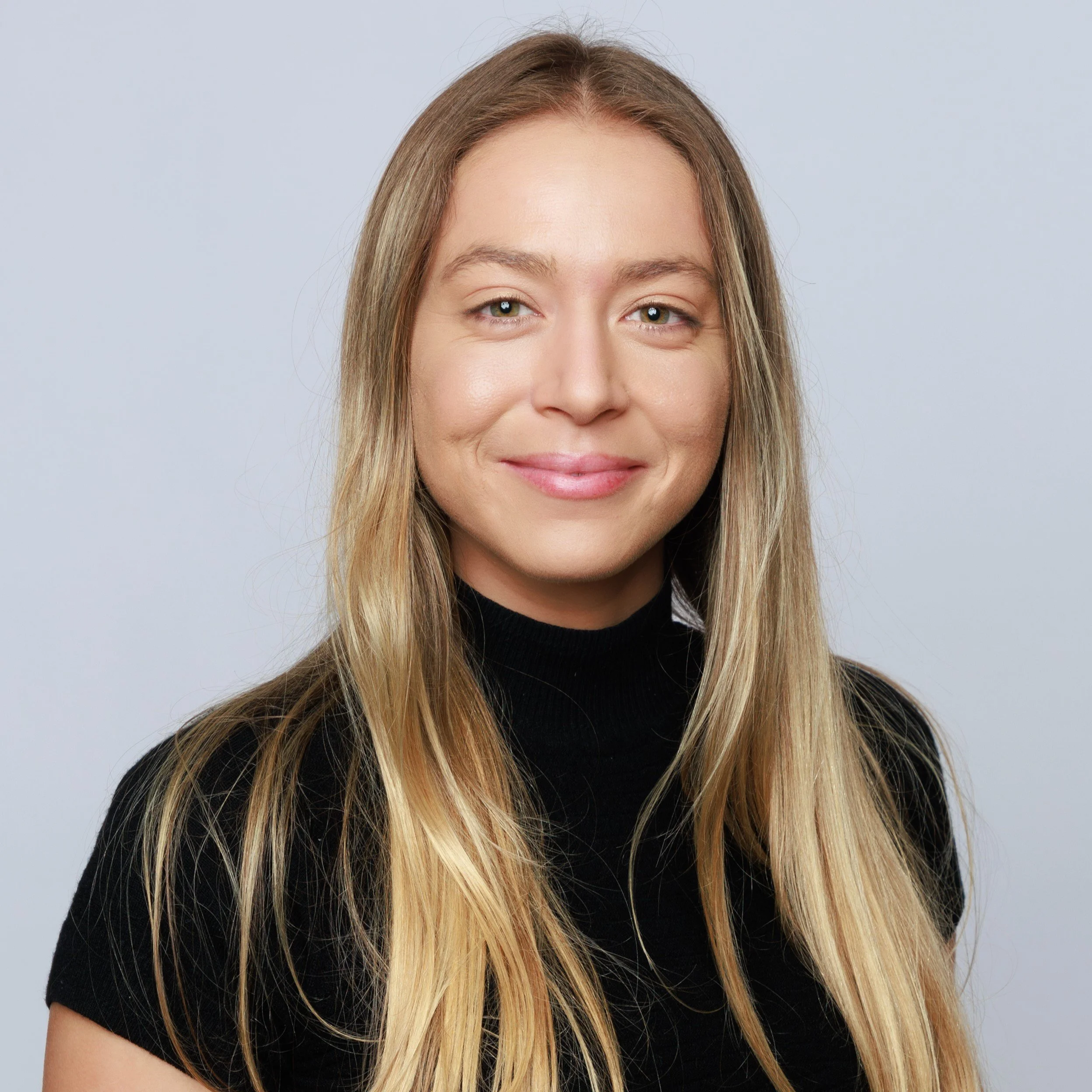 A young woman with long blonde hair, wearing a black turtleneck sweater, smiling against a plain light gray background.