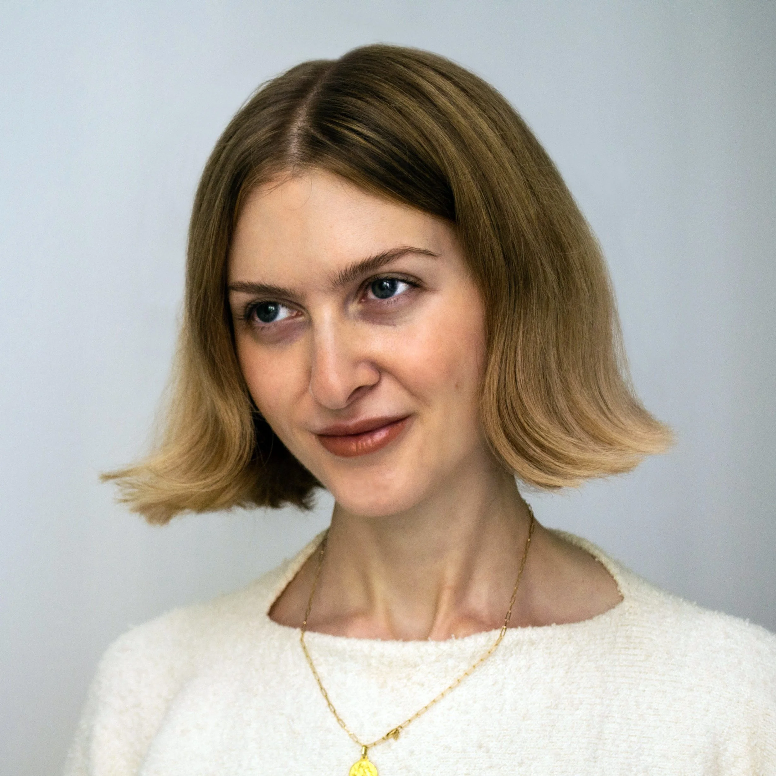 Close-up photo of a woman with shoulder-length blonde hair, wearing a white sweater and a gold necklace, looking to the side with a slight smile.