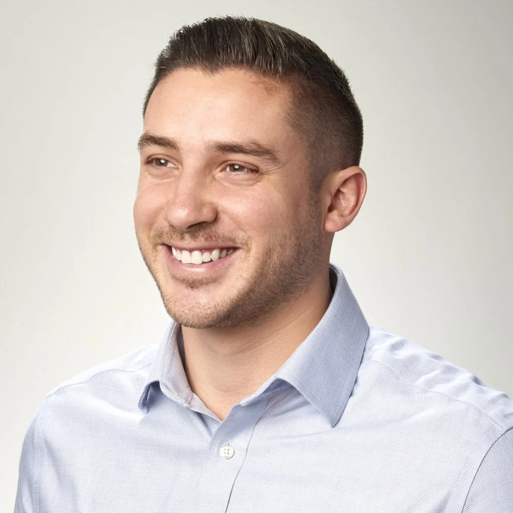 A smiling man with short brown hair, wearing a light gray collared shirt, against a plain light background.