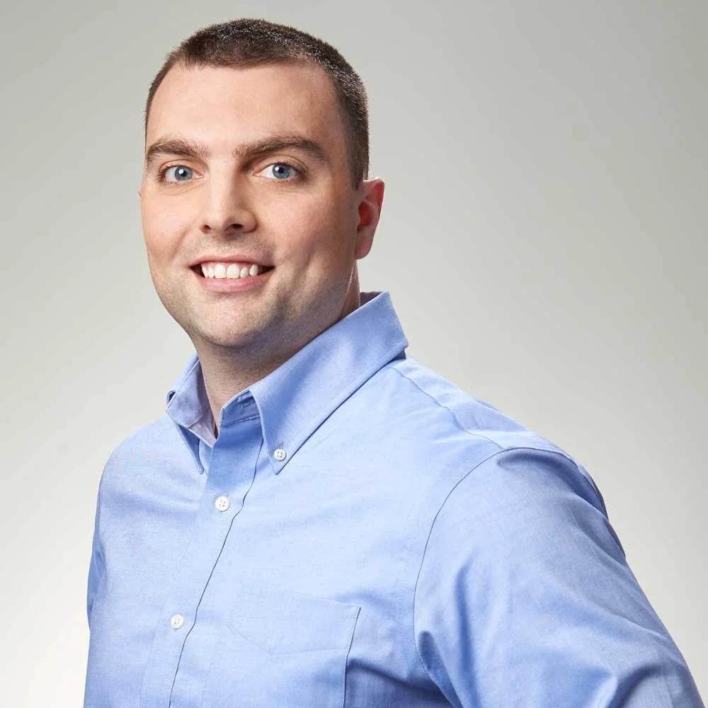 Portrait of a young man with short dark hair, blue eyes, and a friendly smile, wearing a light blue button-down shirt, standing against a plain light gray background.
