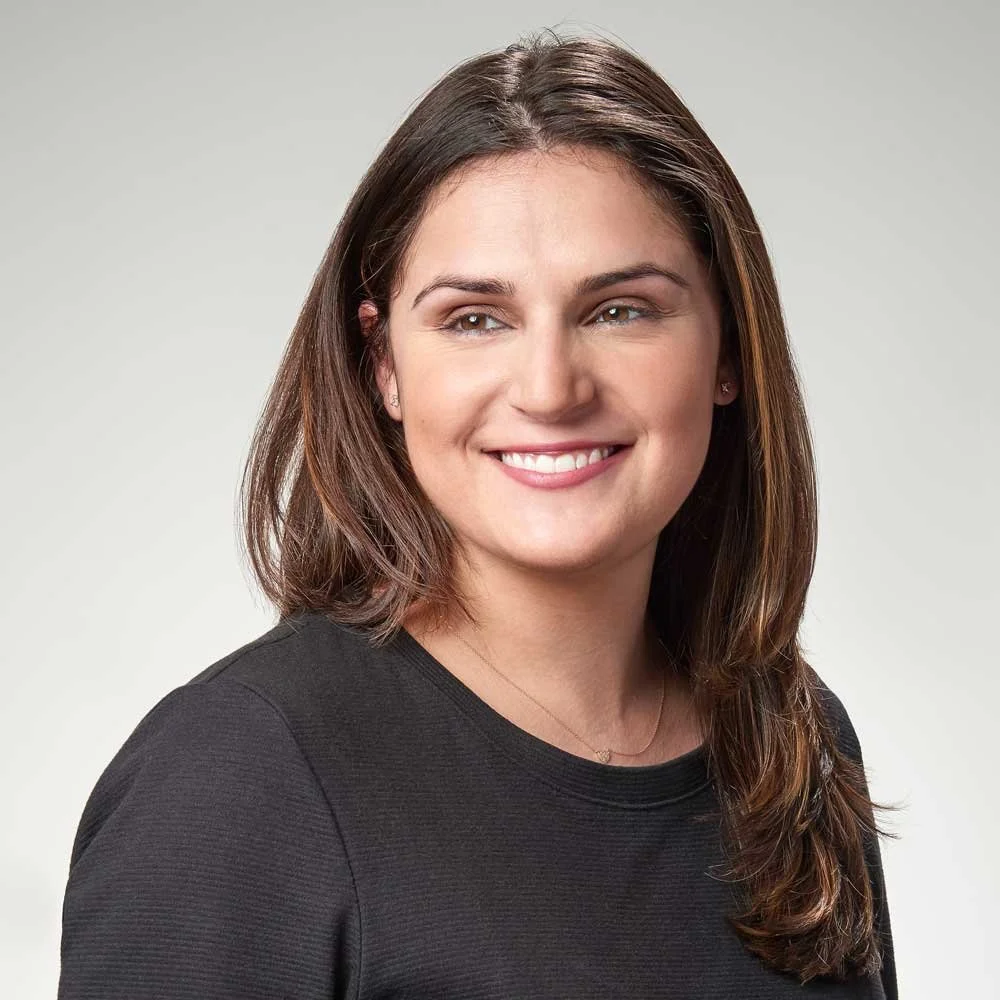 Headshot of a woman with shoulder-length brown hair, smiling, wearing a black top, against a neutral background.