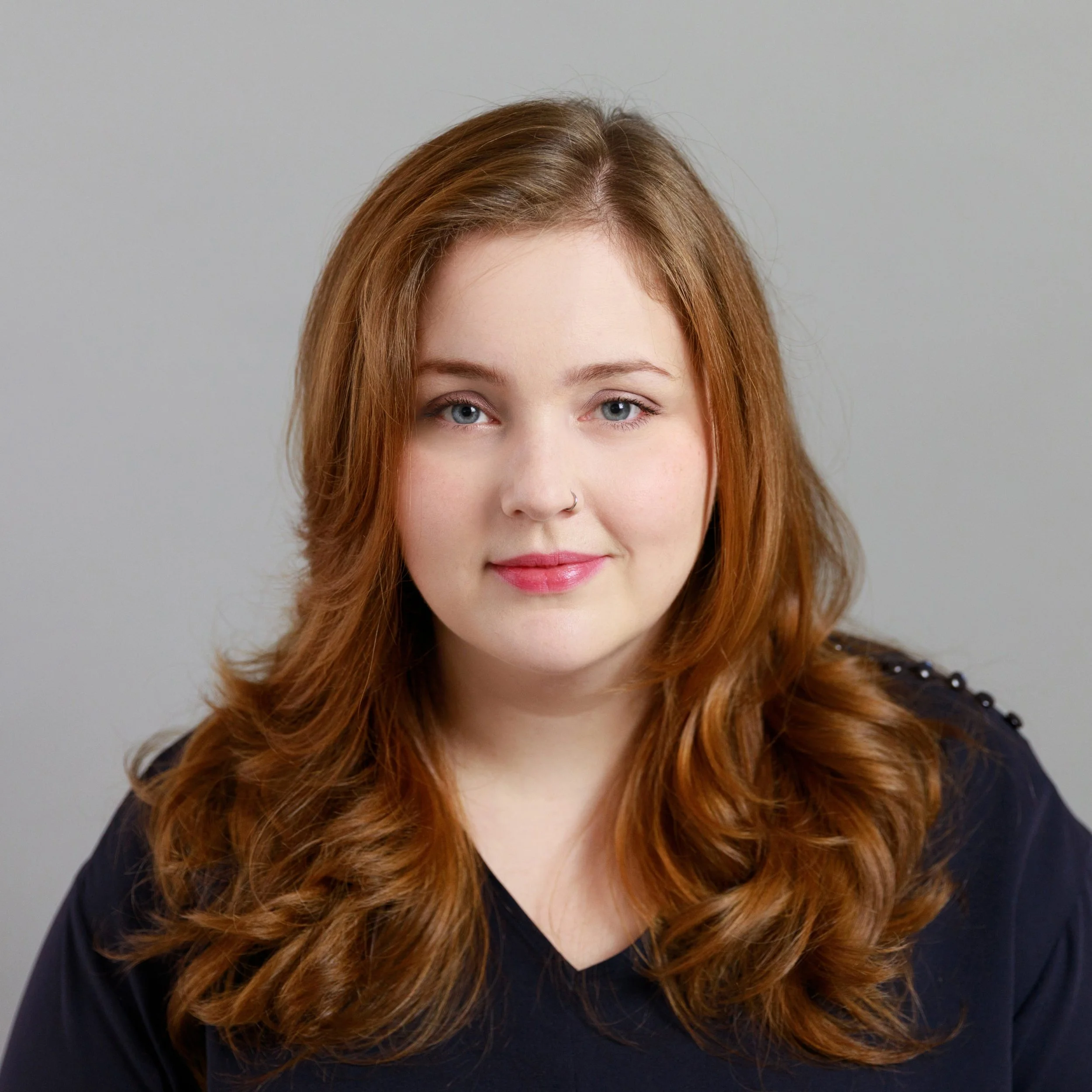 A woman with long wavy red hair, fair skin, blue eyes, wearing a dark top, slightly smiling, against a plain gray background.
