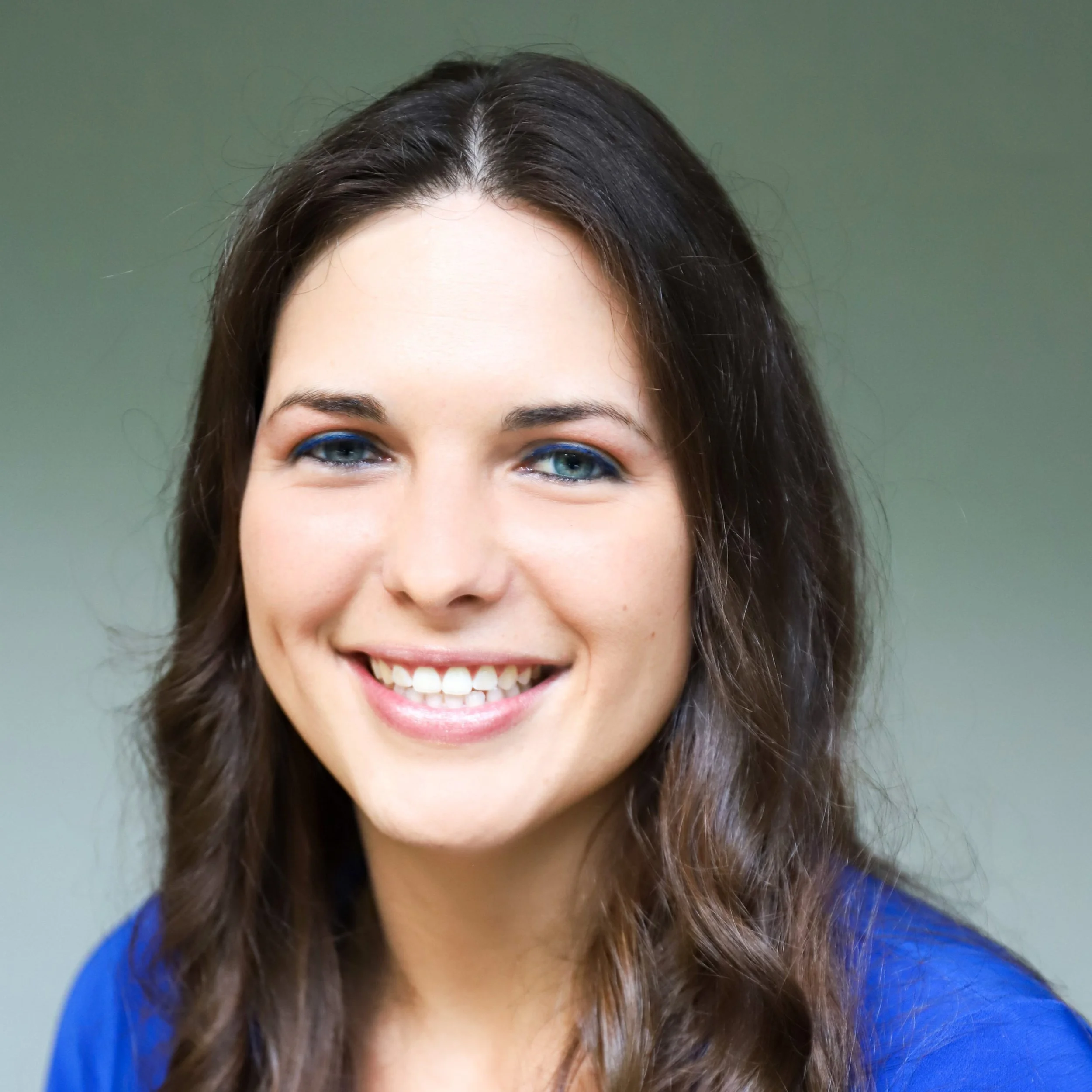 A smiling woman with long brown hair, blue eyes, wearing a blue top, in front of a plain, light green background.