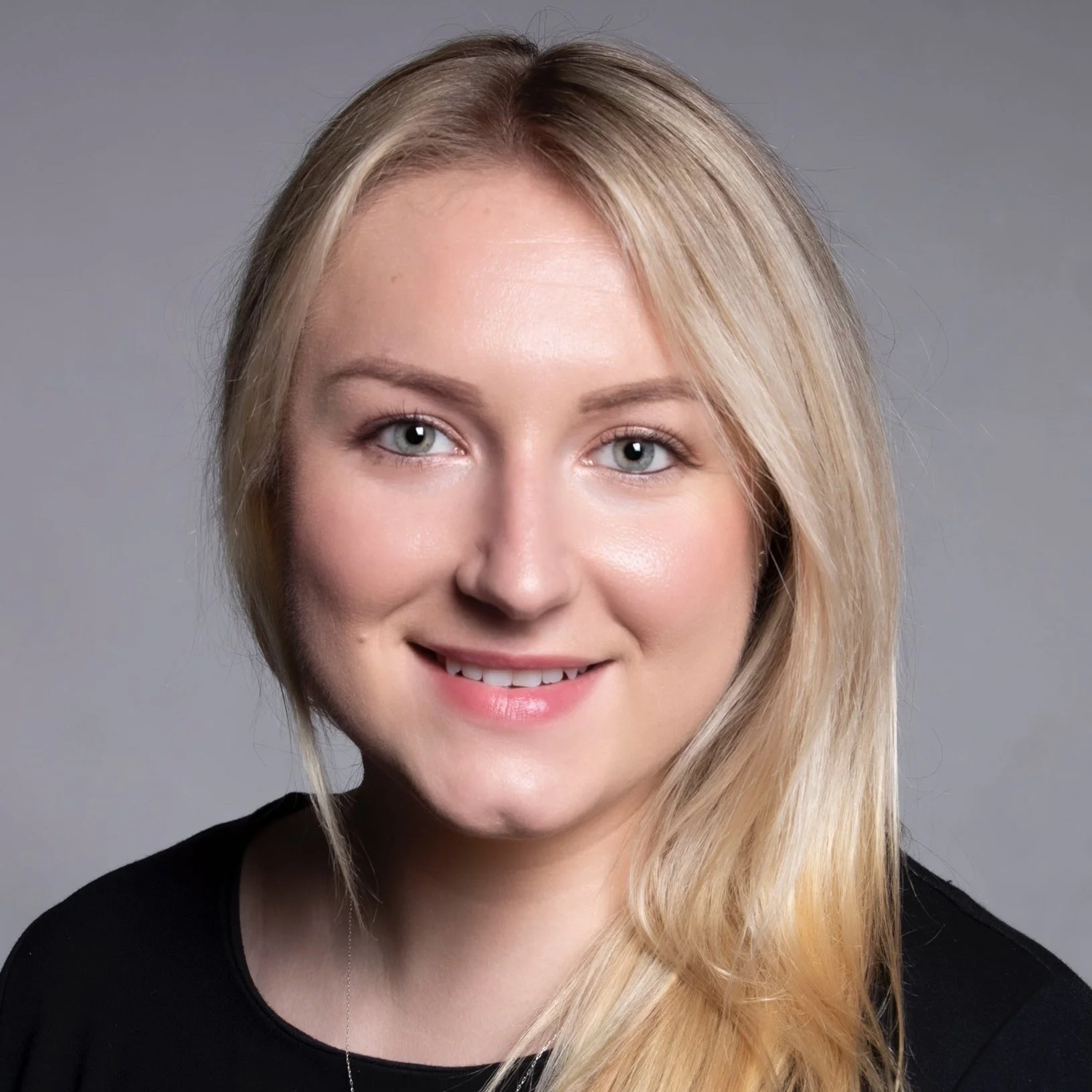 Headshot of a young woman with blonde hair, blue eyes, and fair skin, smiling, wearing a black top, against a gray background.