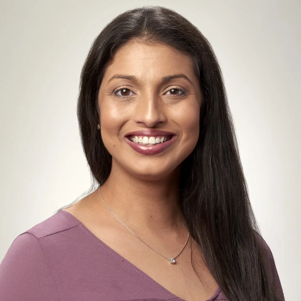 A woman with long dark hair smiling, wearing a purple top and a silver necklace, against a neutral background.