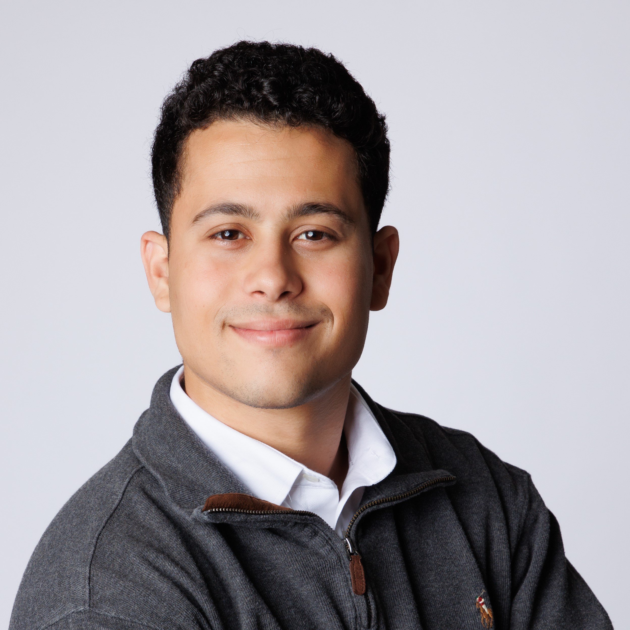 Headshot of a young man with short dark hair, wearing a white shirt and a dark gray zip-up sweater, smiling gently against a plain light gray background.