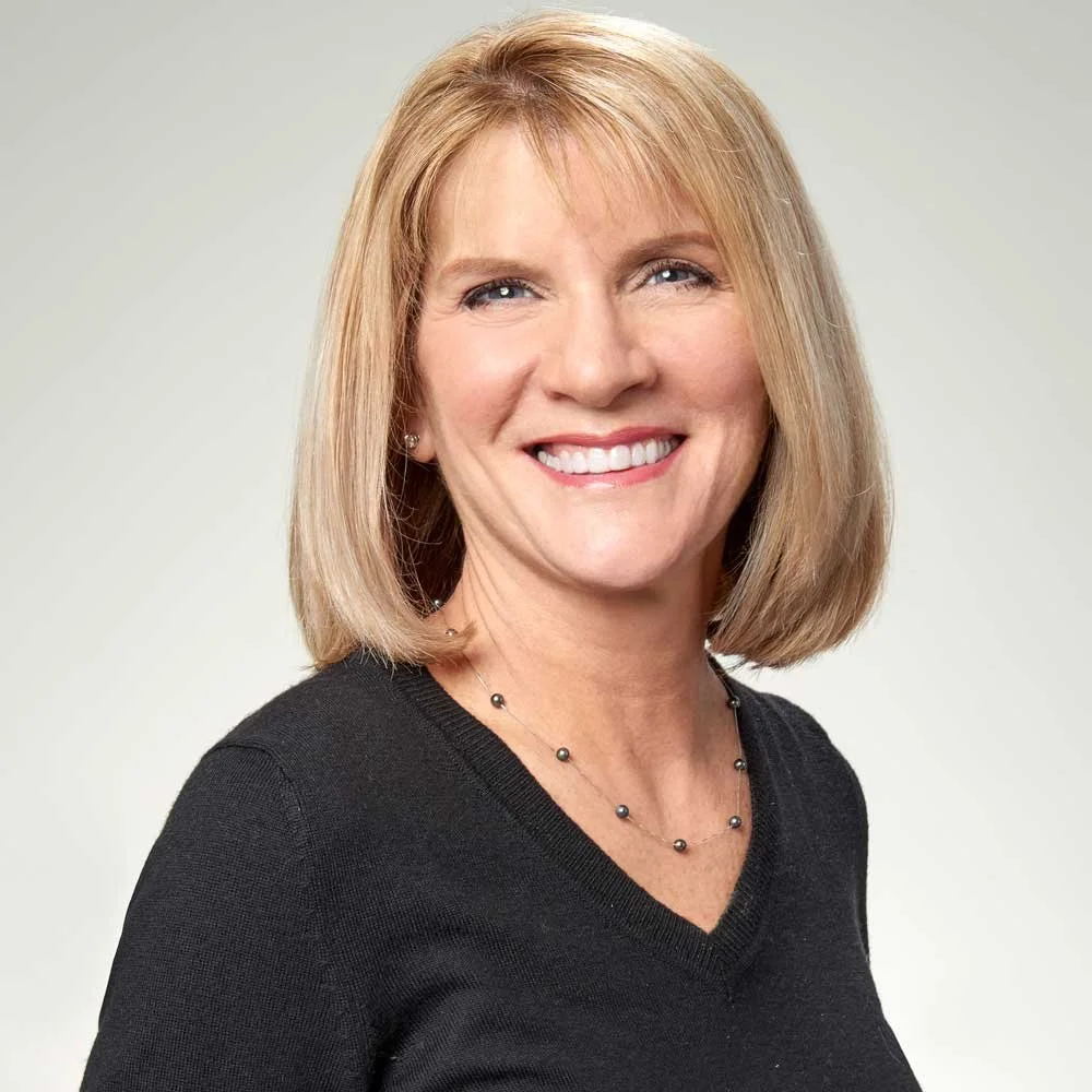 A smiling woman with shoulder-length blonde hair wearing a black top and a beaded necklace.